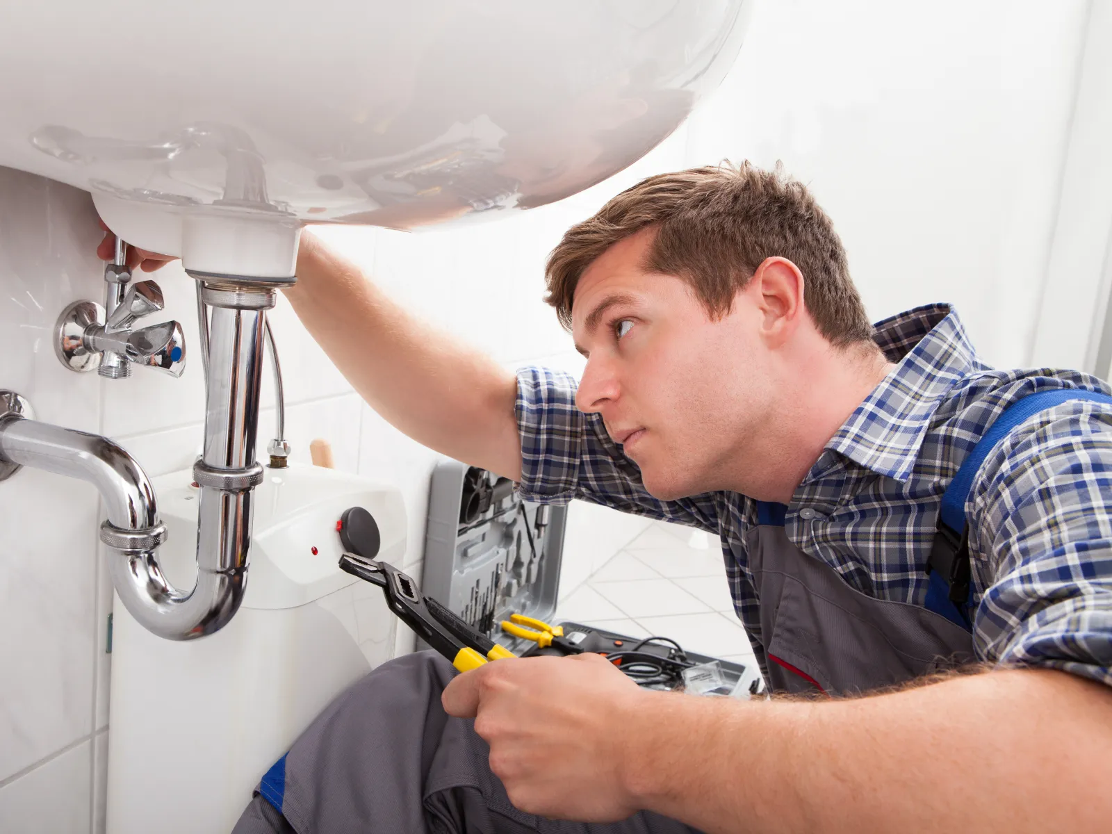Plumber in checkered shirt fixing bathroom sink plumbing with wrench under white ceramic sink.