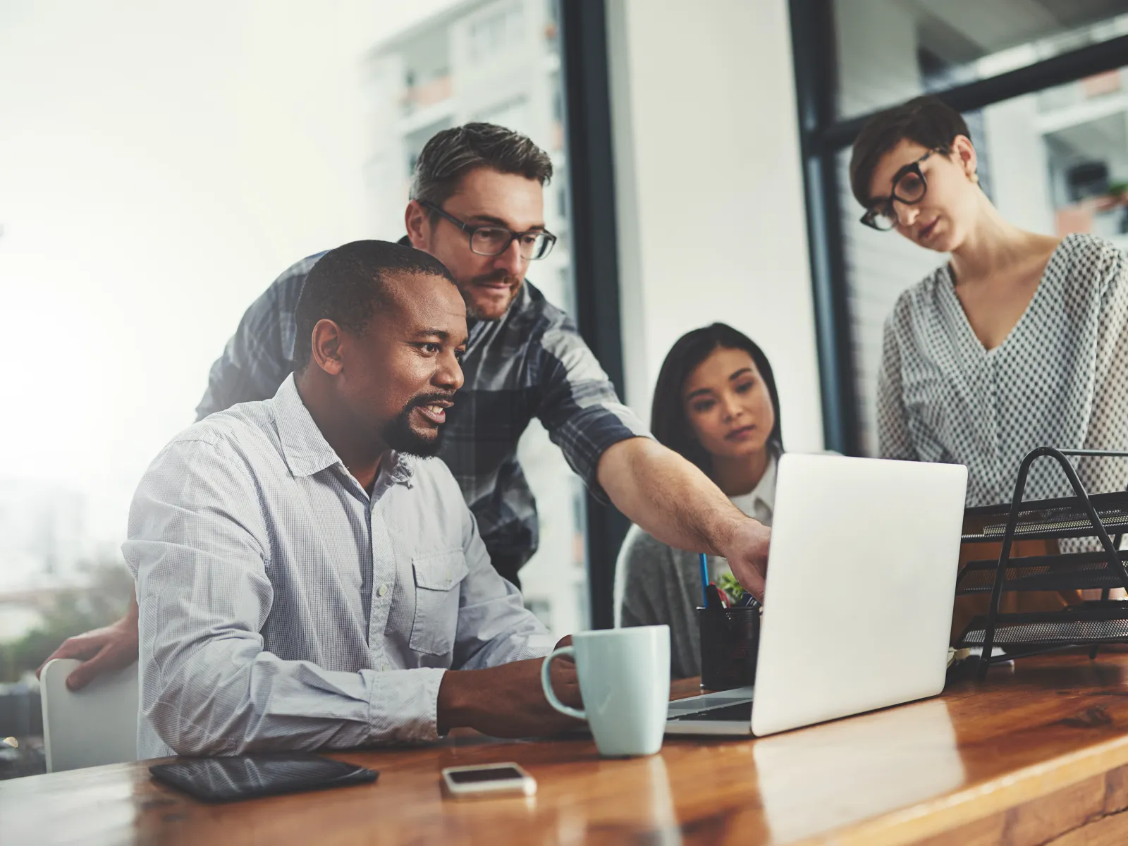Diverse team collaborating around a laptop in a modern office with natural light and wooden table.