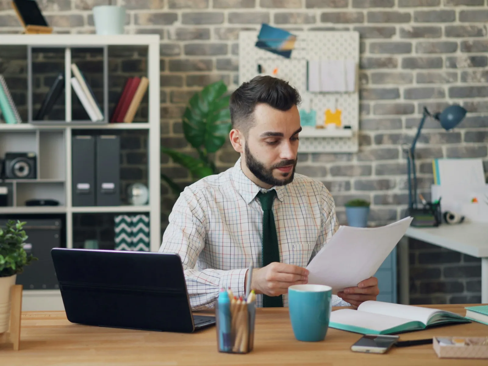 Man in office reading papers with laptop, books, coffee cup, and plants on desk under brick wall.