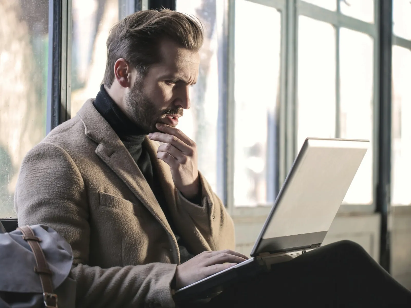 Man in beige coat focused on laptop while sitting near large windows in bright room