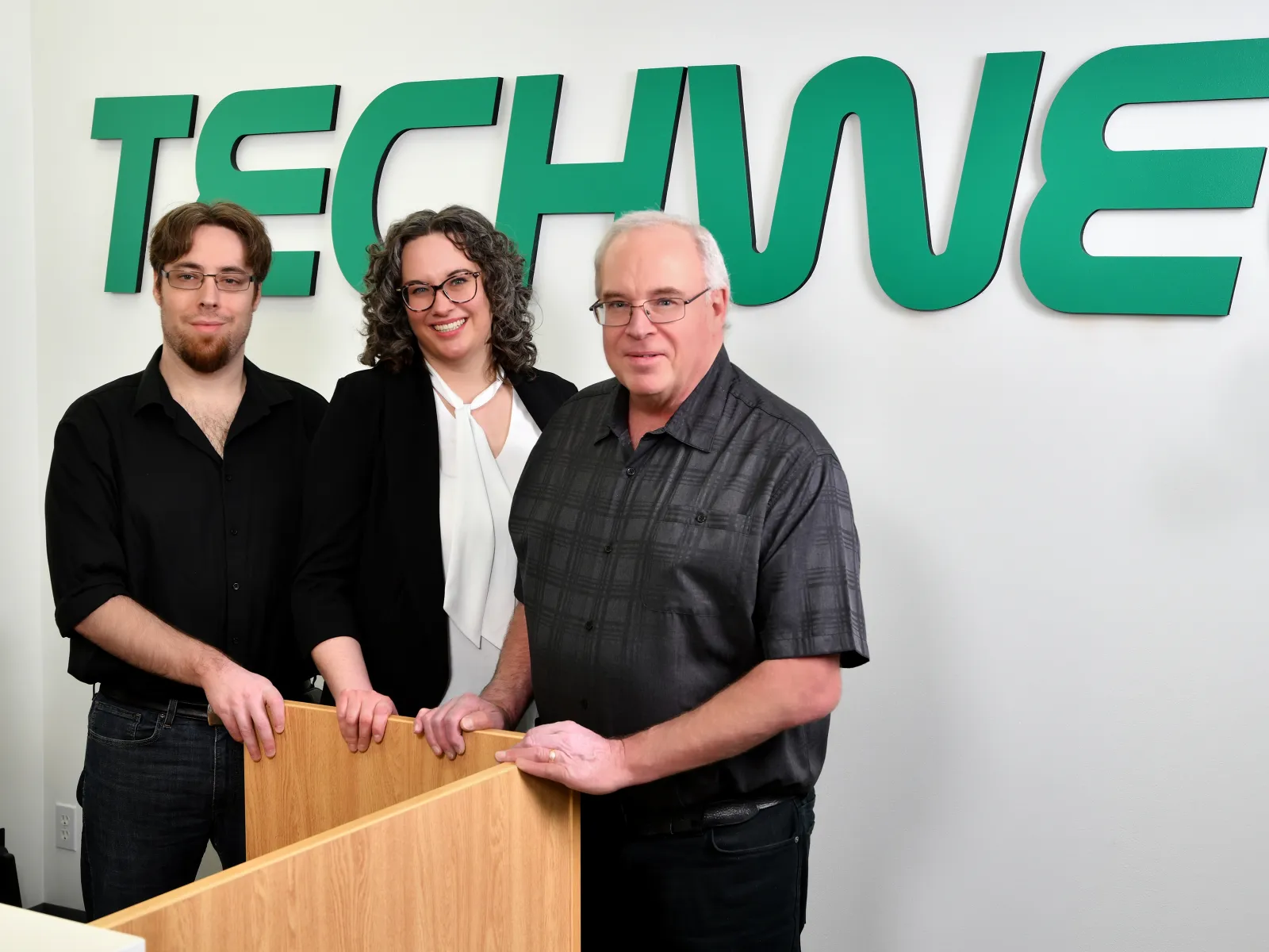 Three professionals standing together in front of a TechWell sign, smiling confidently in office setting.