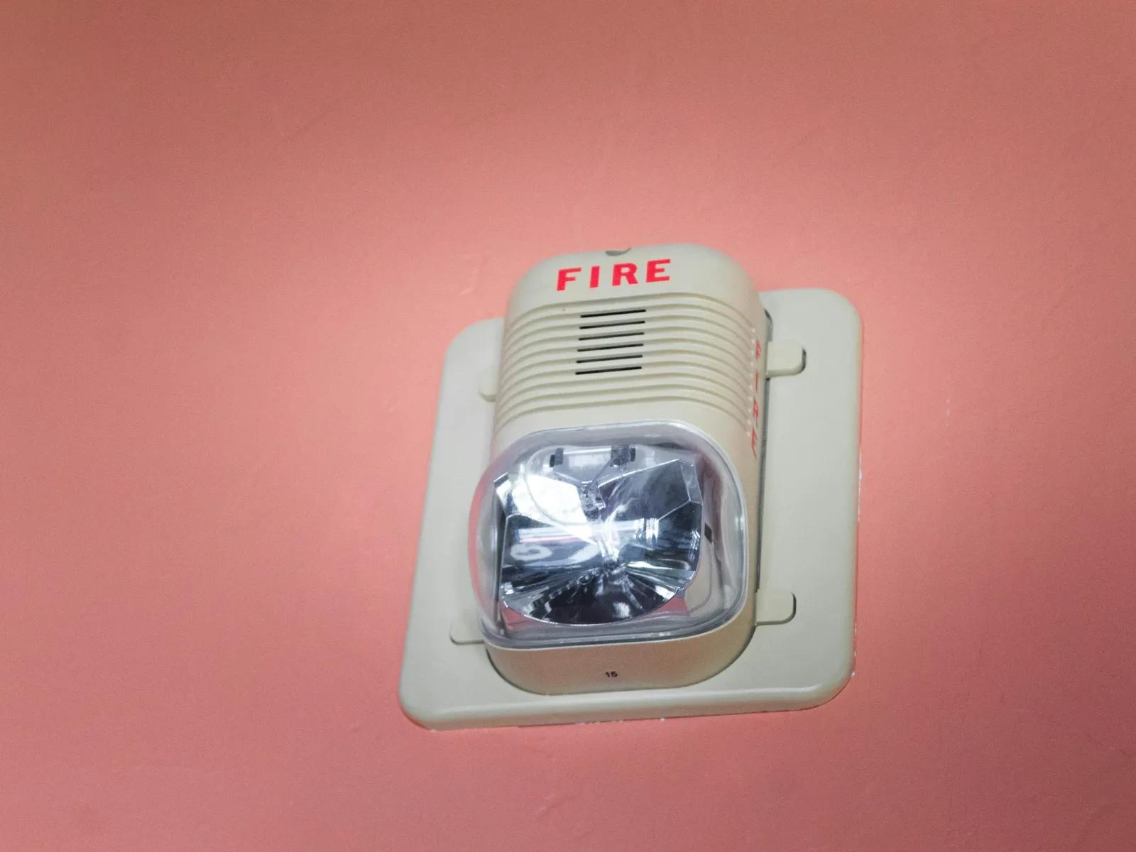 Wall-mounted fire alarm strobe light on a pink surface with red FIRE text and white casing.