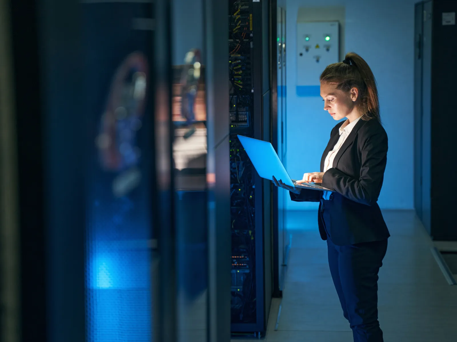 Woman in business attire working on a laptop in a dimly lit server room with blue lighting.