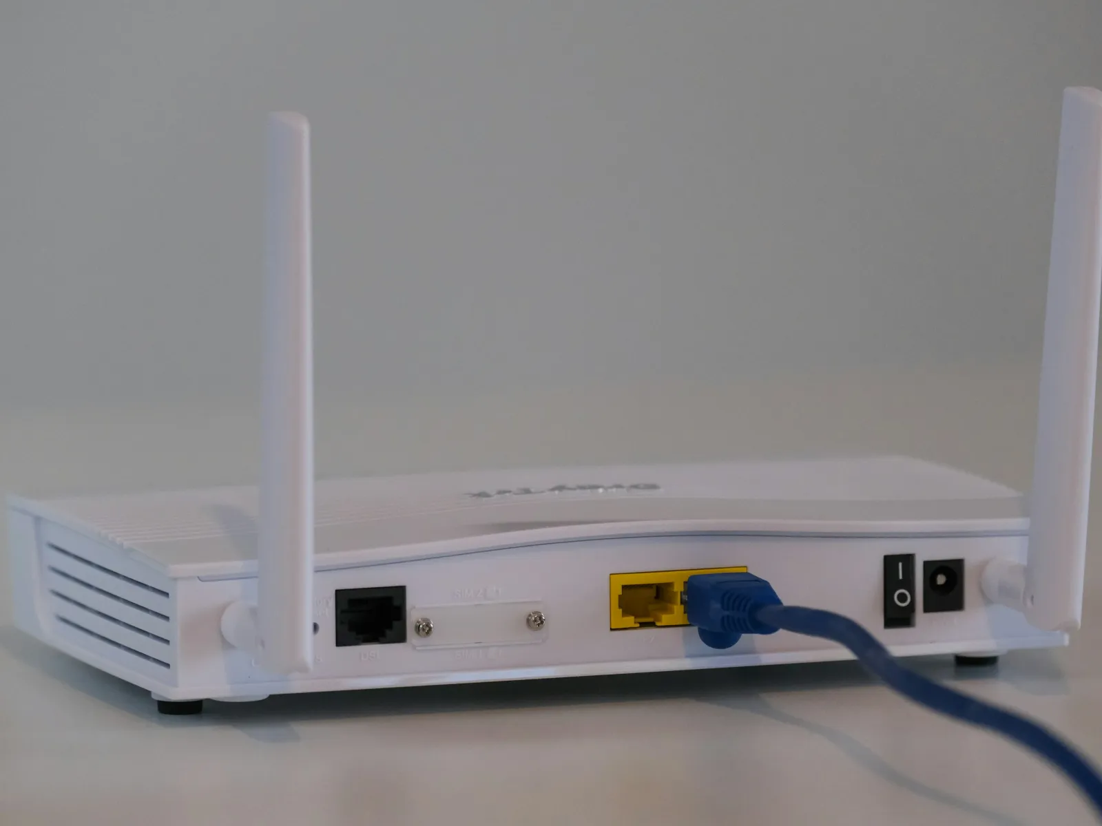 White wireless router with two antennas and a blue Ethernet cable plugged into a yellow port on a gray surface.