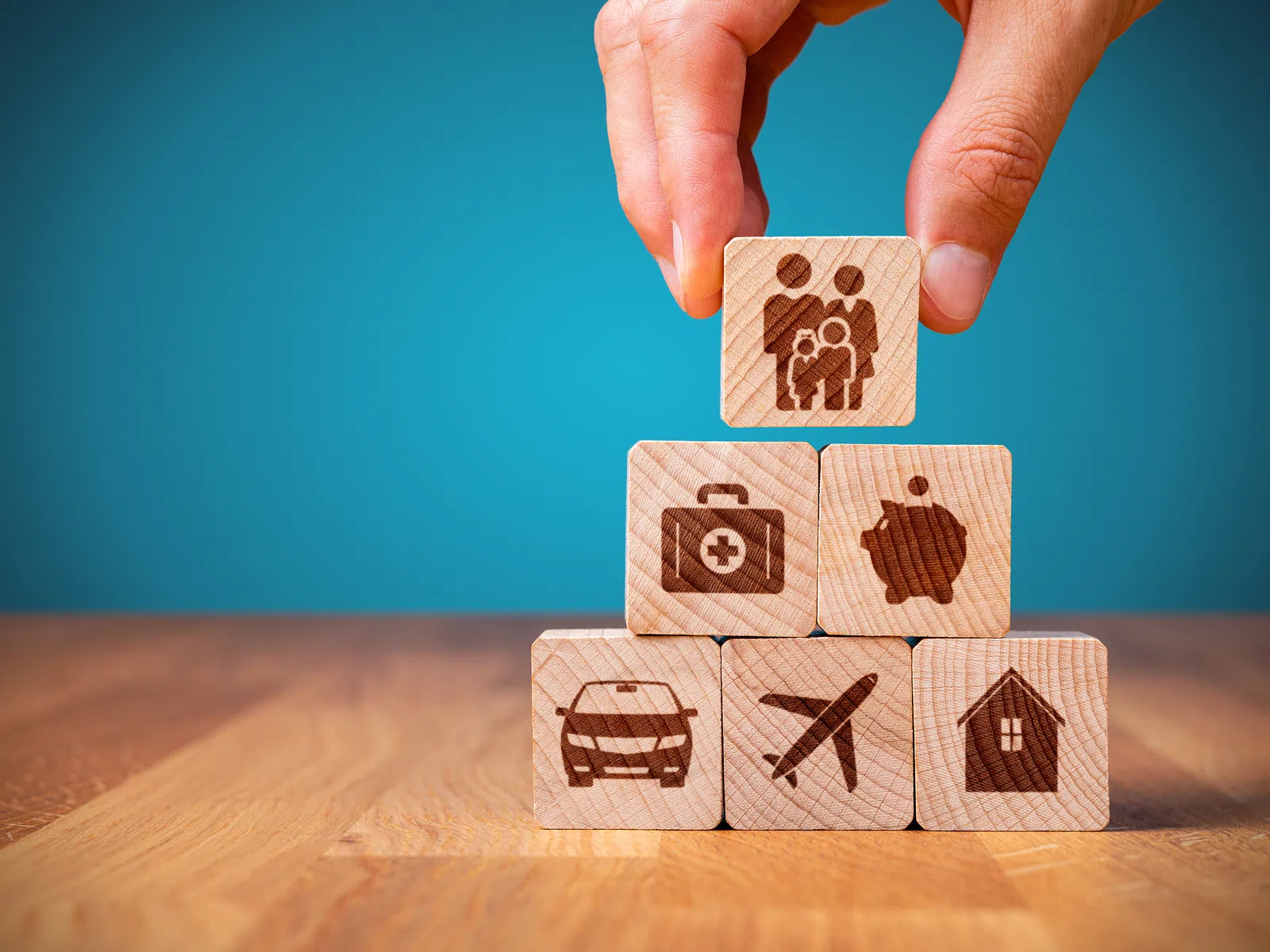 Hand stacking wooden blocks with icons representing family, health, savings, car, travel, and home on a wooden surface.