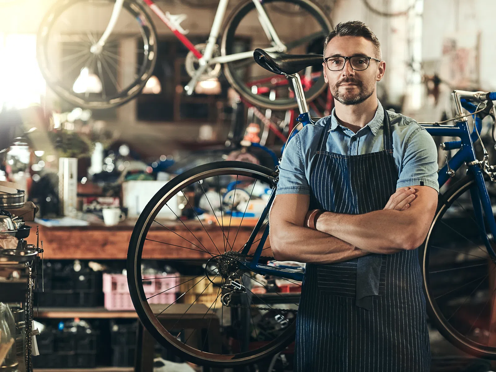 Male bicycle mechanic with arms crossed standing next to bike in a workshop filled with tools and bike parts