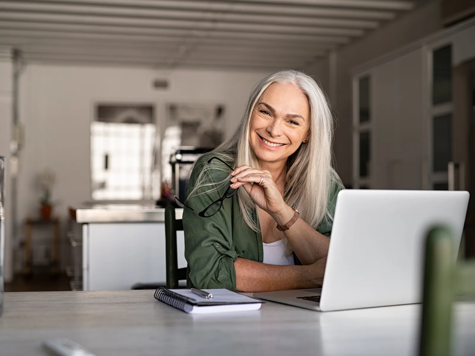 Smiling mature woman with gray hair working on laptop at home with notepad and glasses in hand