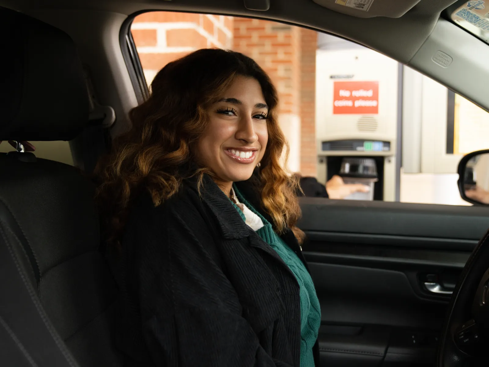 Happy woman sitting in car using a drive-thru ATM machine with brick wall background