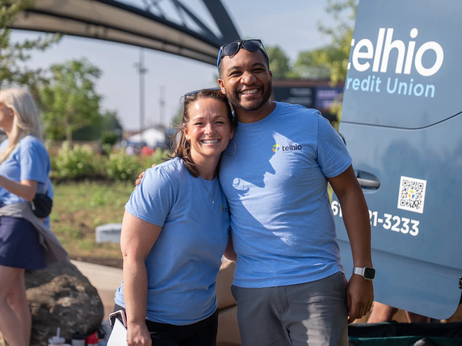 Two smiling people wearing matching blue Telhio Credit Union shirts standing outside near a branded van.