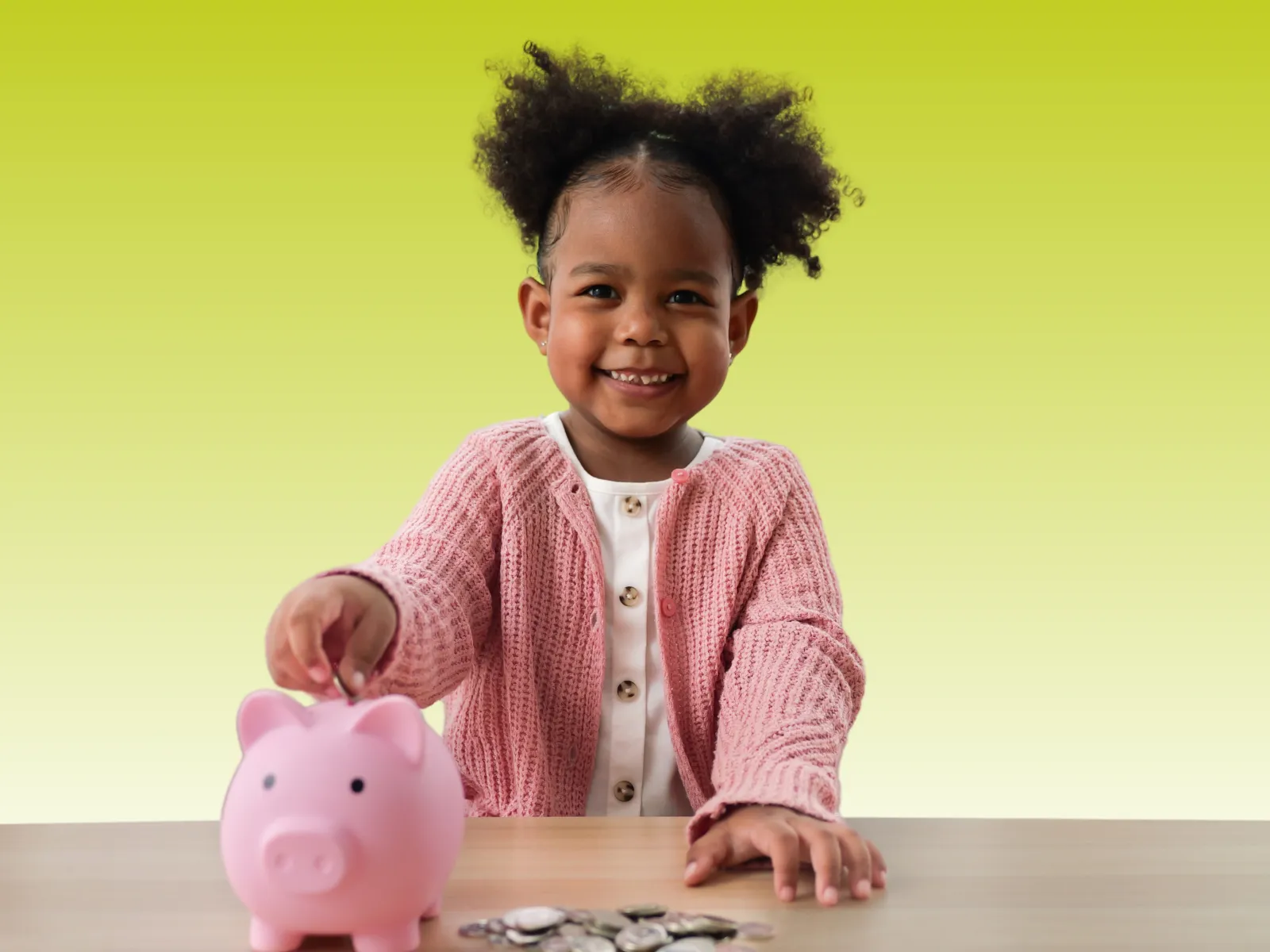 Smiling young girl in pink sweater putting coin into pink piggy bank on wooden table