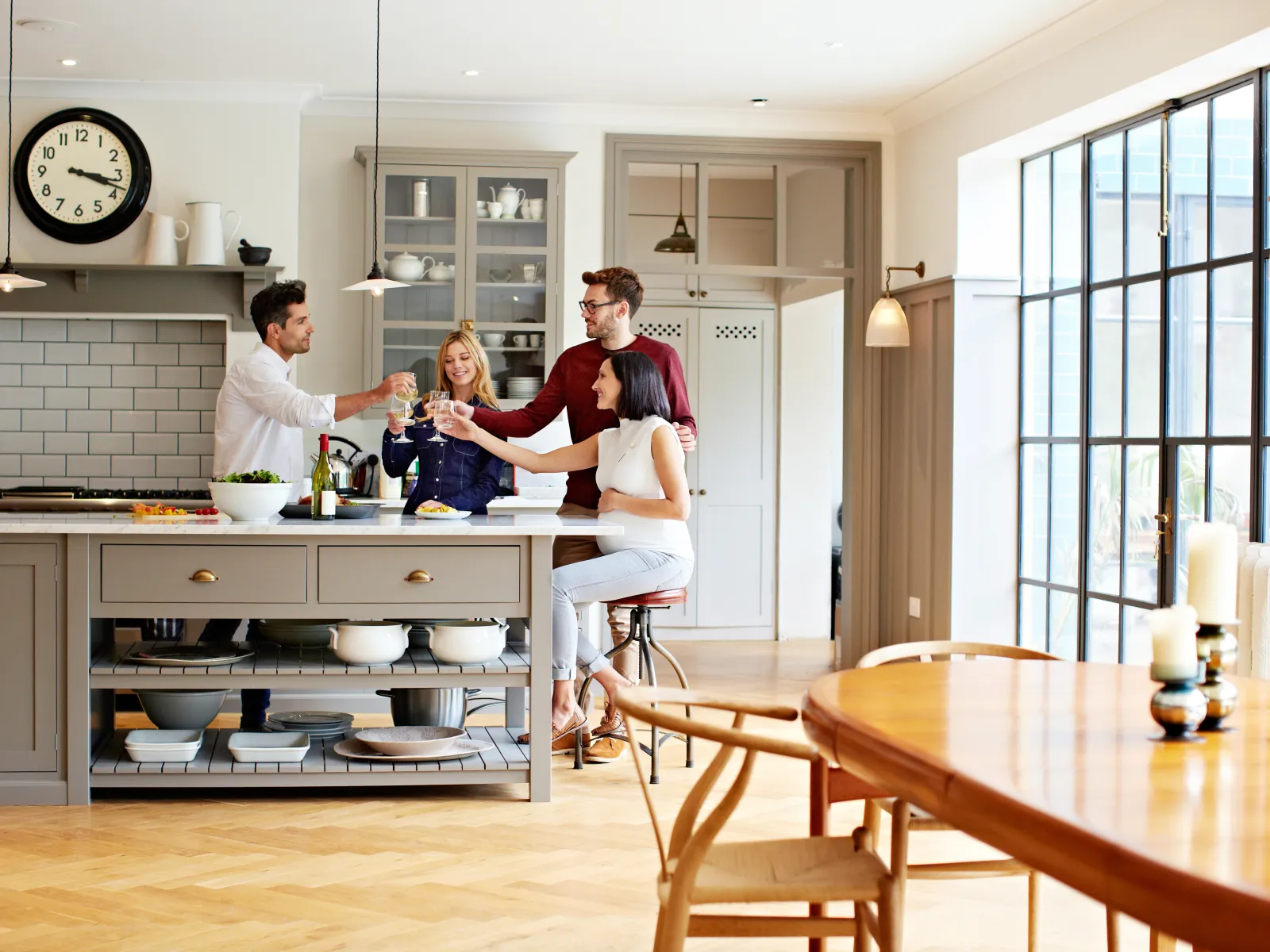 Four friends toasting drinks together in a modern kitchen with large windows and wooden floors.