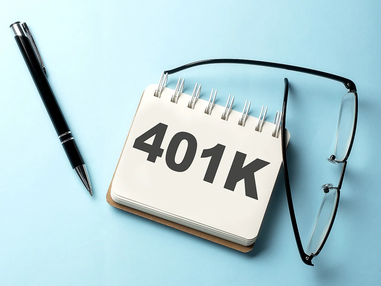 Spiral notebook displaying 401K text with a pen and eyeglasses on light blue background
