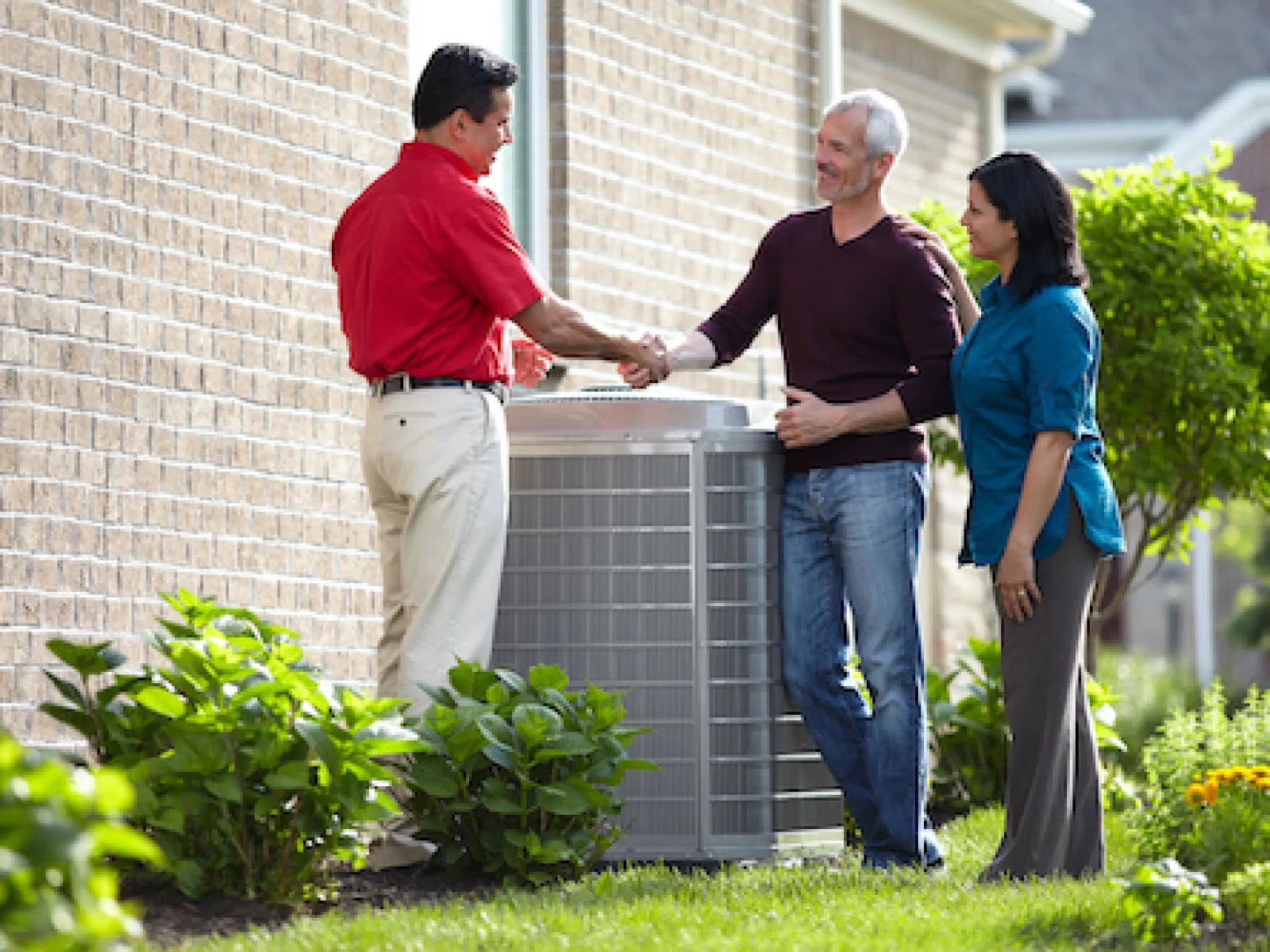 Technician shaking hands with couple outside near air conditioning unit by brick house on sunny day