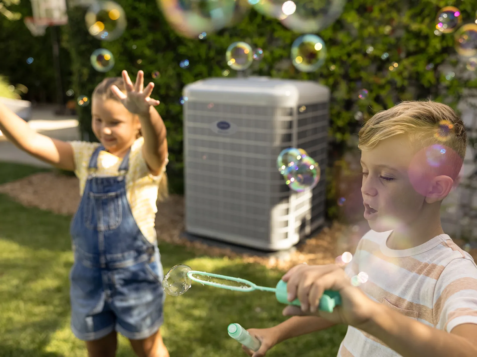 Two children playing with bubbles in a sunny backyard near an outdoor air conditioning unit.