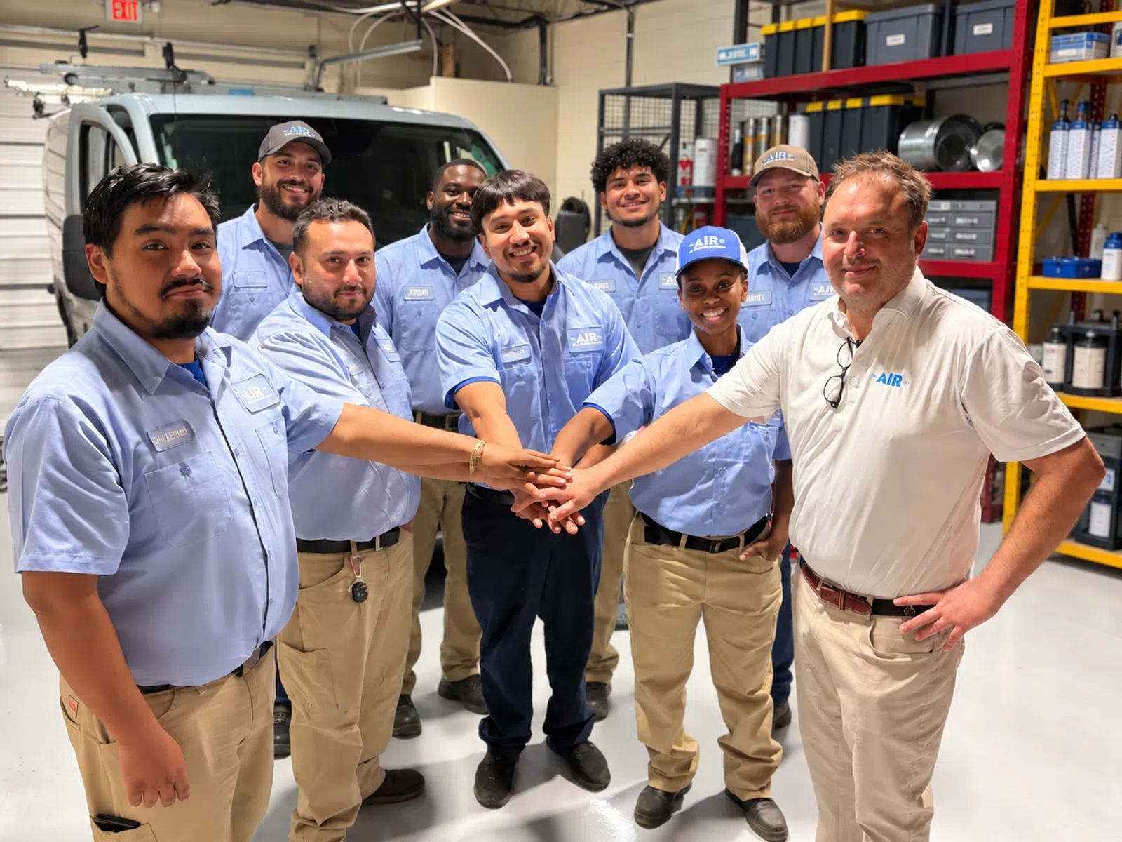 Team of friendly HVAC technicians wearing light blue uniforms joining hands in a workshop with equipment and van.