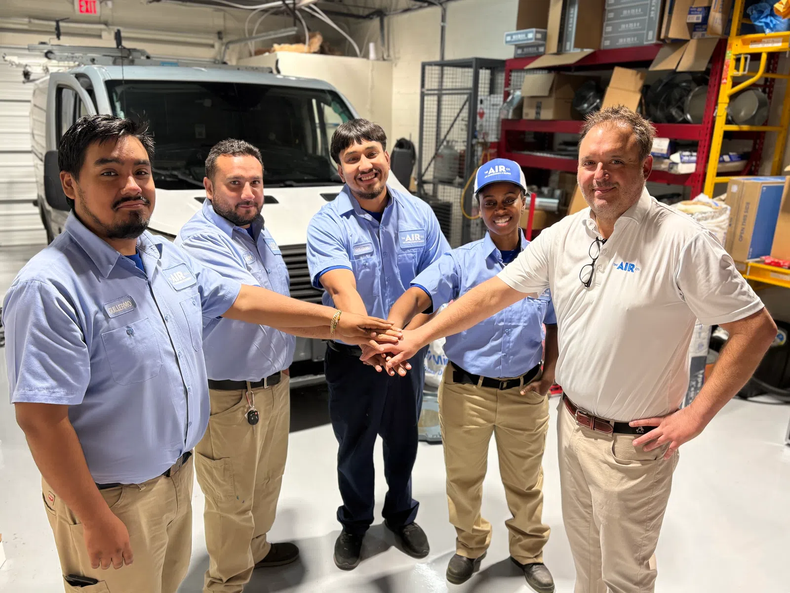 Five HVAC technicians standing in a workshop, united with hands stacked, wearing blue and white AIR uniform shirts.