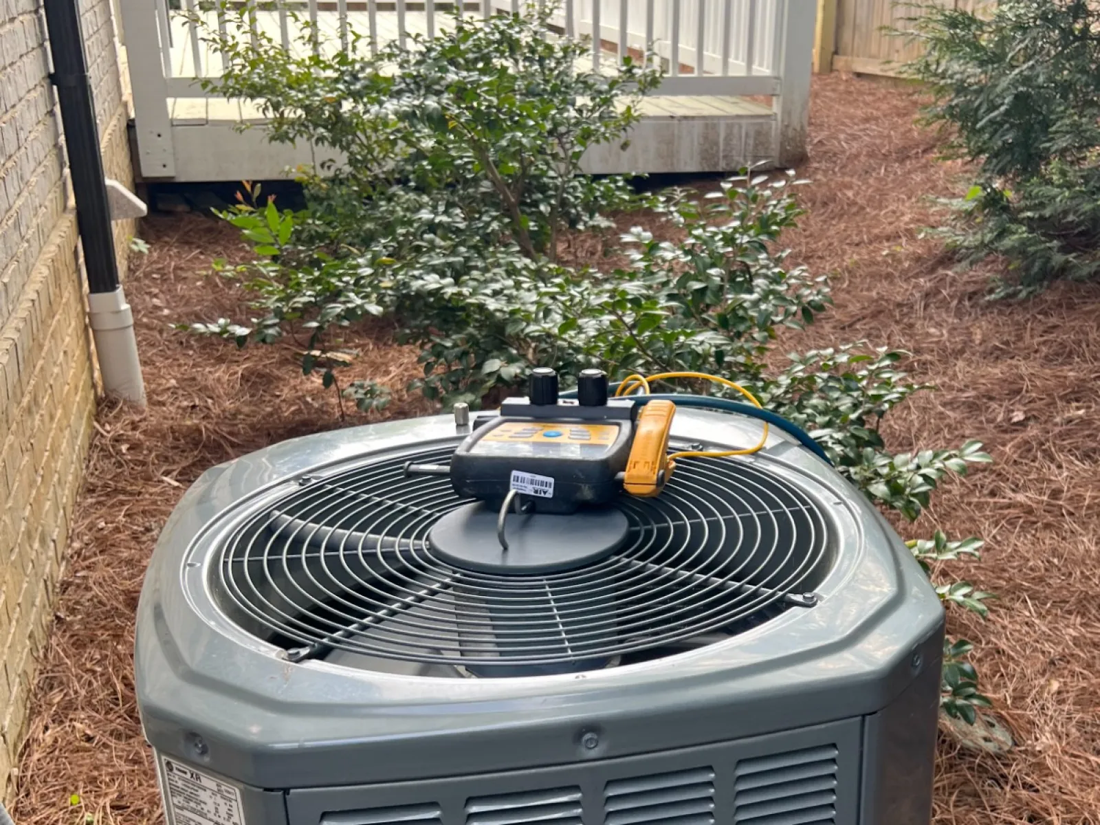 Outdoor air conditioning unit near brick wall with diagnostic tools on top surrounded by pine straw and bushes.