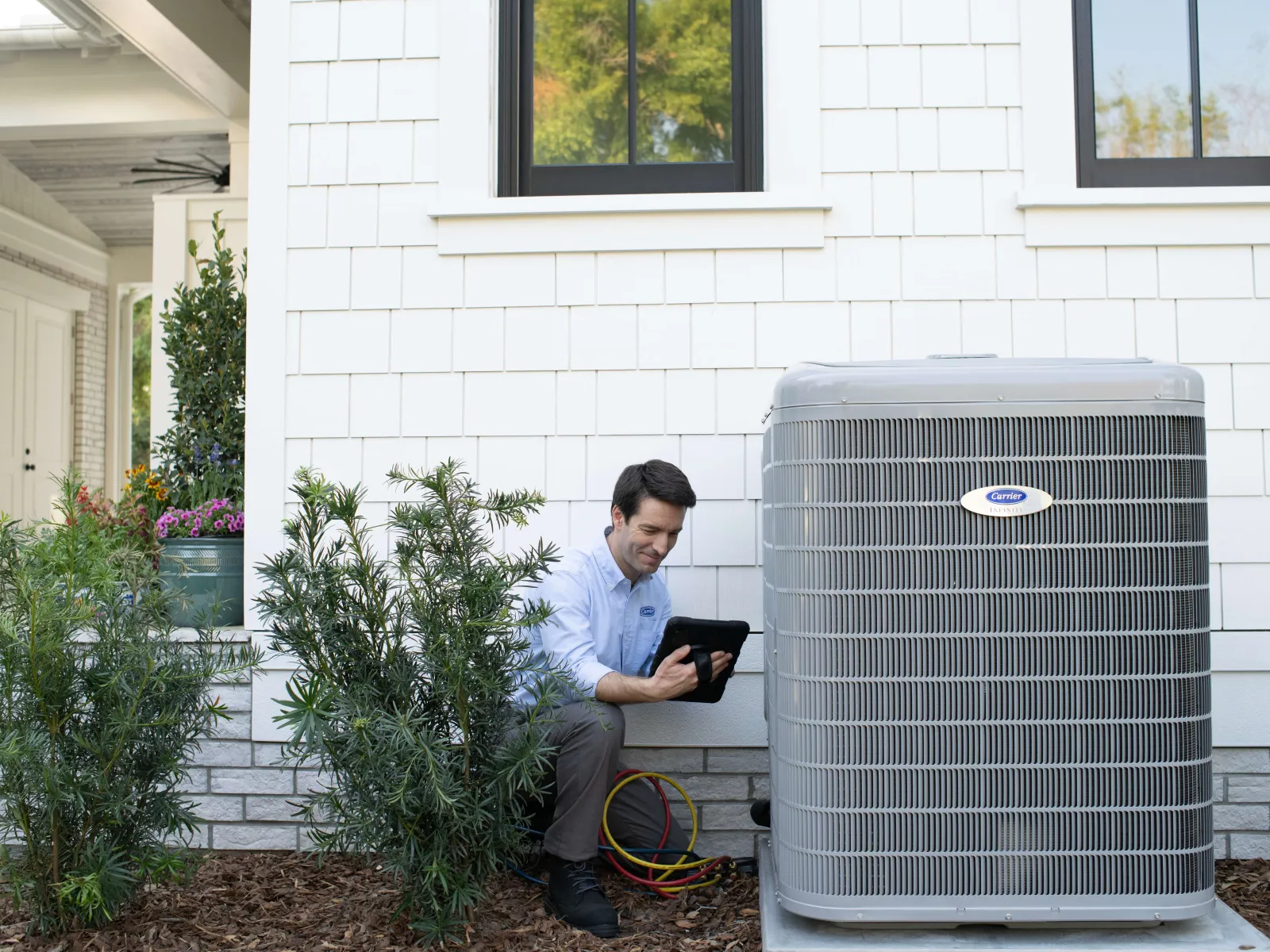 Technician inspecting and working on a home air conditioning unit outside a white house during daytime.
