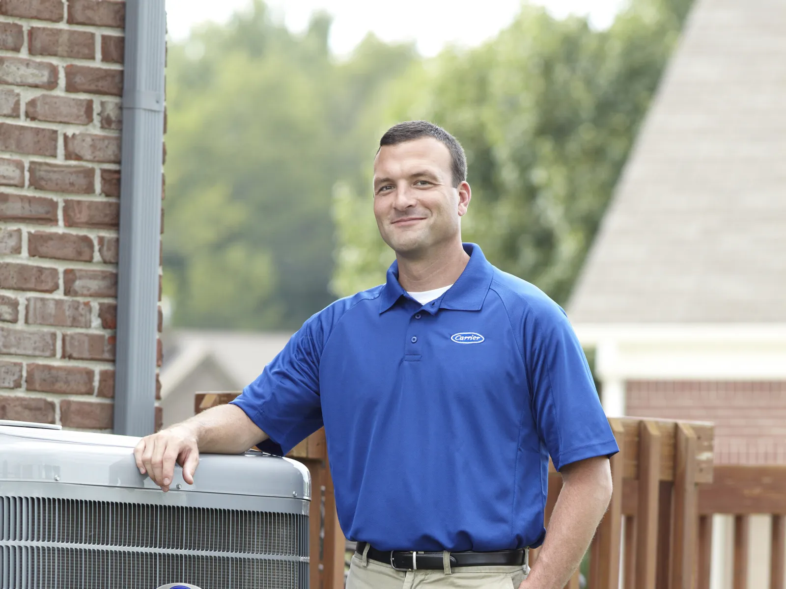 Technician in blue shirt standing next to a Carrier HVAC unit outside a home with greenery and wooden fence.