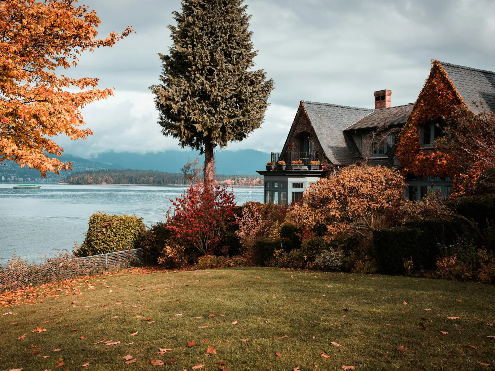 Cozy lakeside house surrounded by autumn trees and colorful foliage under a cloudy sky.