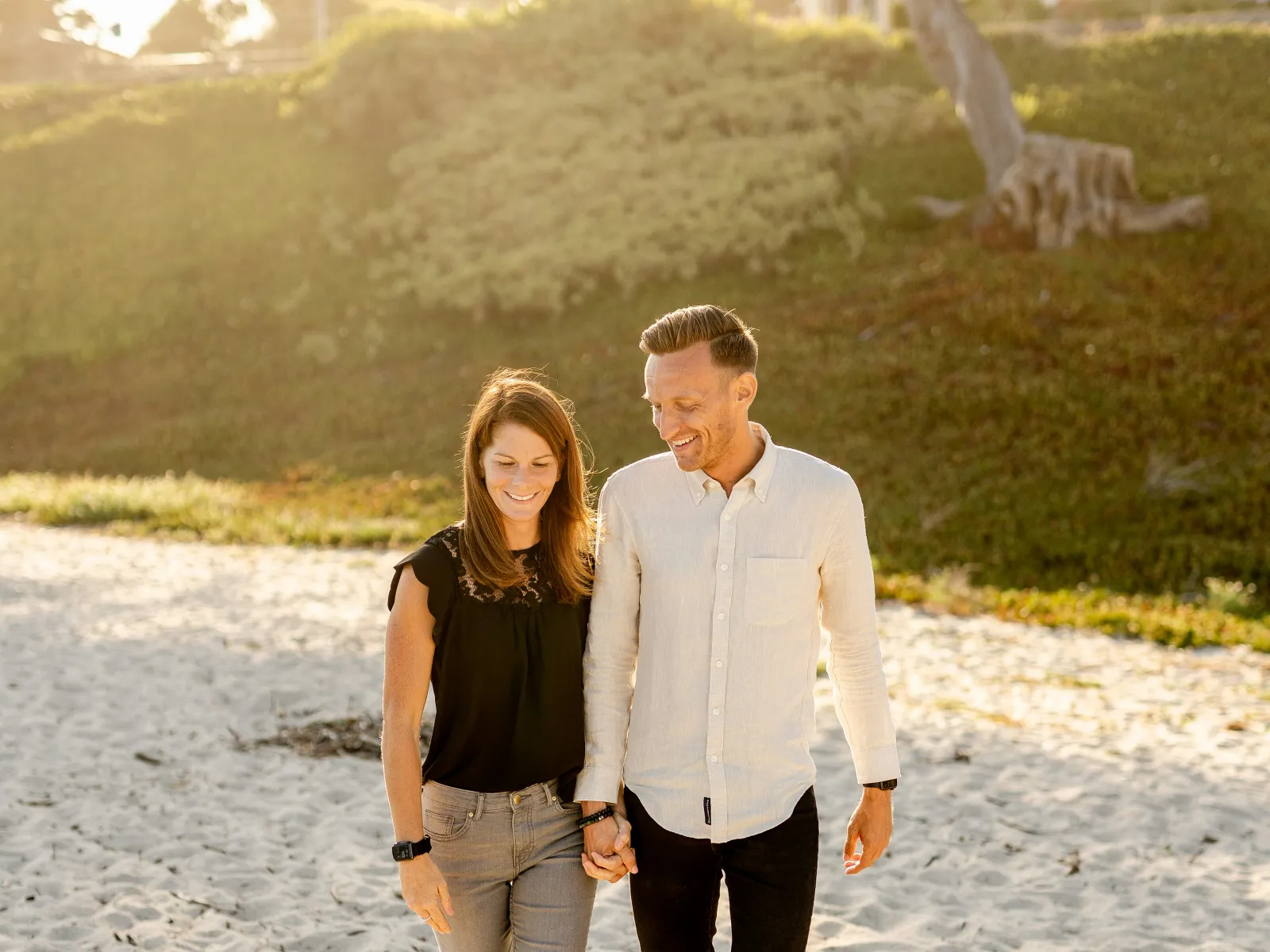 Couple holding hands and walking barefoot on sandy beach during sunny day with greenery and house behind