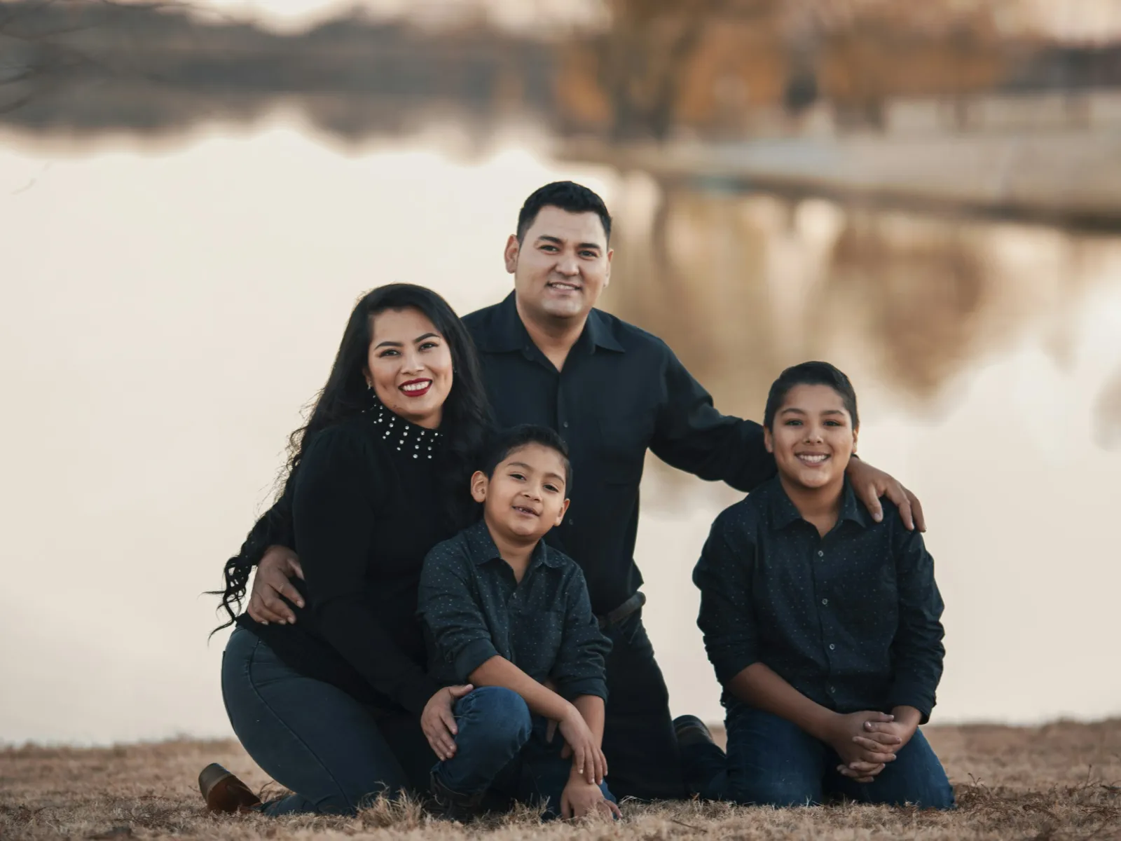 Happy family of four dressed in dark casual clothes posing by a calm lake with autumn foliage background.