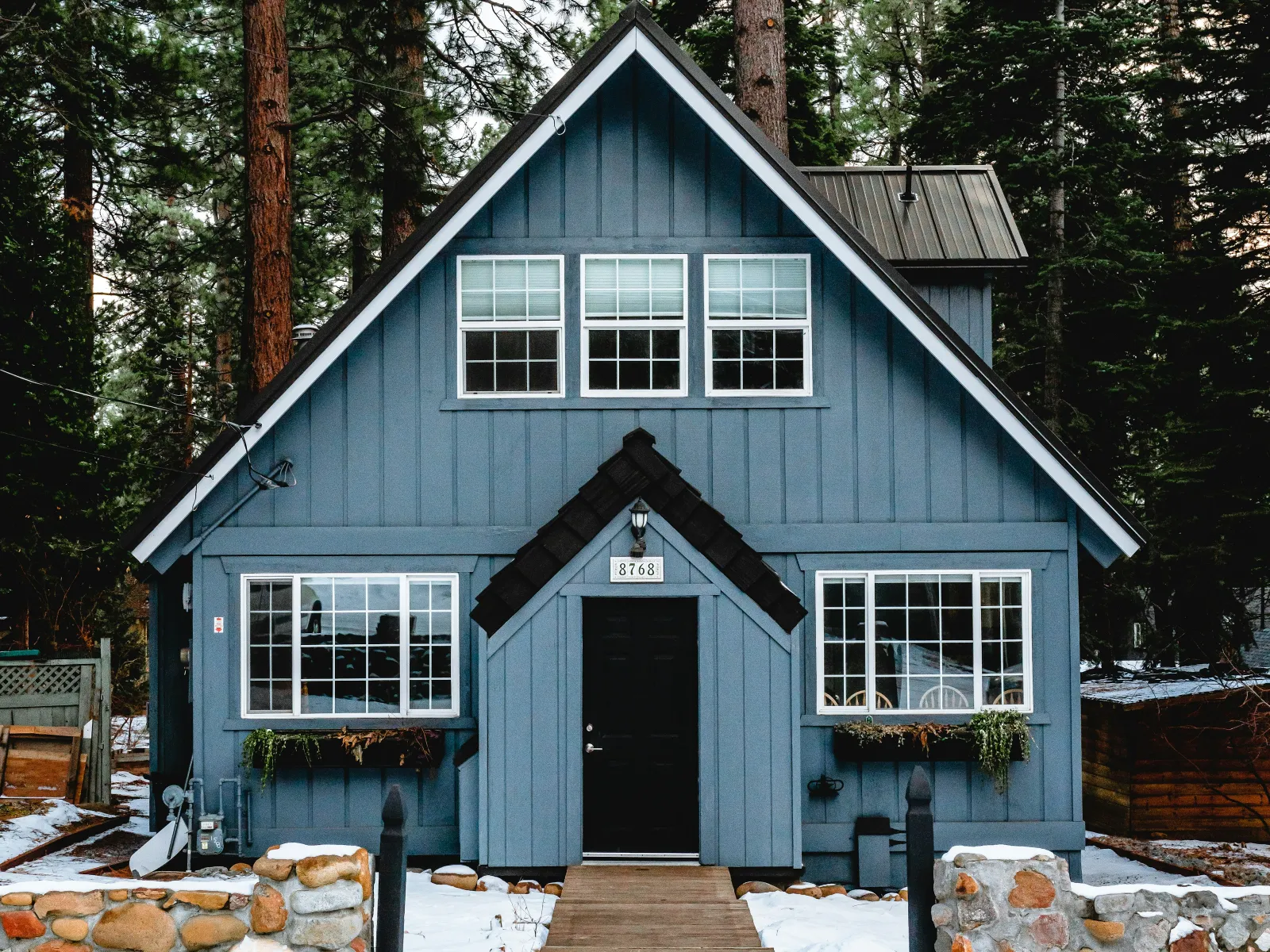Blue cabin house with black door surrounded by tall pine trees and snow-covered ground in winter.