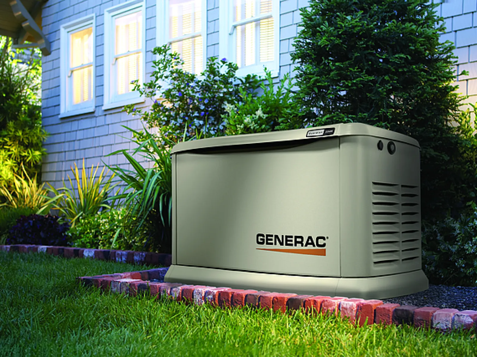 Generac standby generator installed on a brick platform beside a house surrounded by green plants and grass.