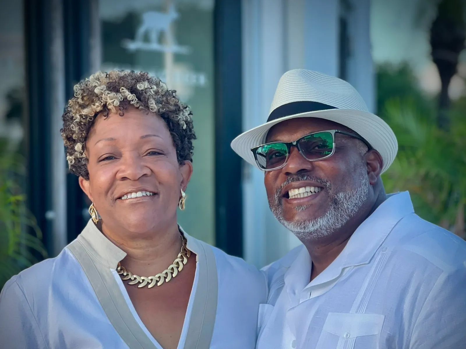 Smiling mature couple wearing white attire, man with hat and glasses, woman with curly hair and gold necklace.