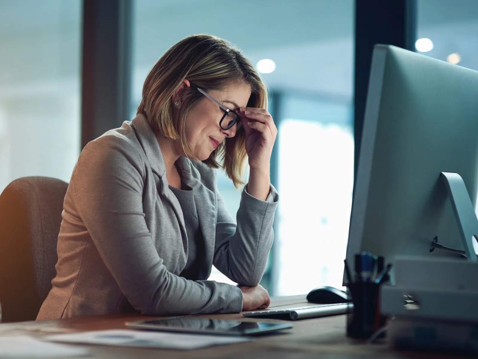 Stressed businesswoman in glasses sitting at desk working on desktop computer in a modern office.