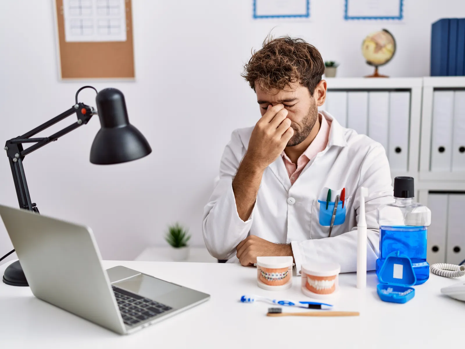 Tired male dentist in white coat sitting at desk with dental models, laptop, toothbrush, and mouthwash in office.