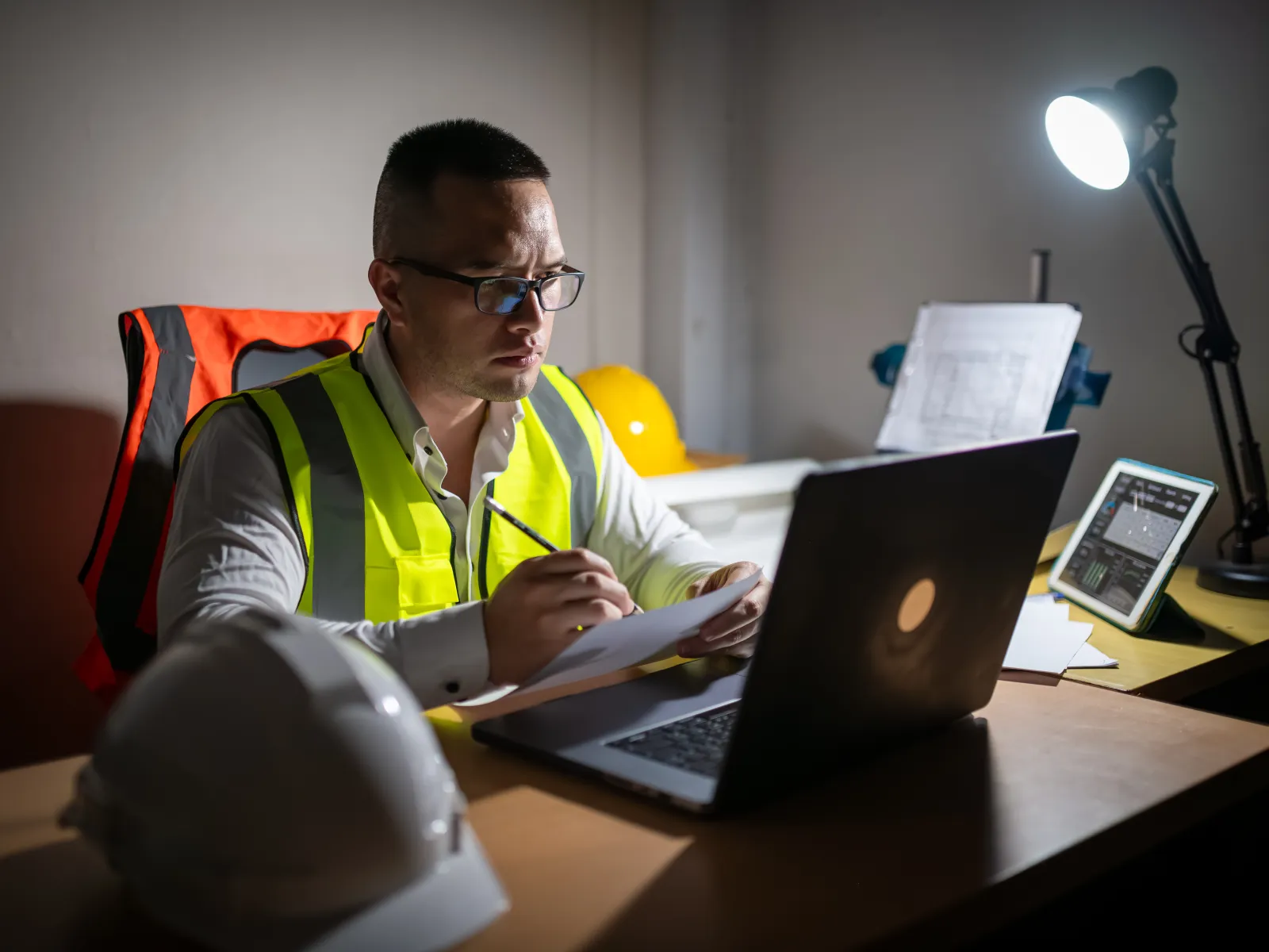 Engineer in a safety vest working late at desk with laptop and documents, illuminated by desk lamp indoors.