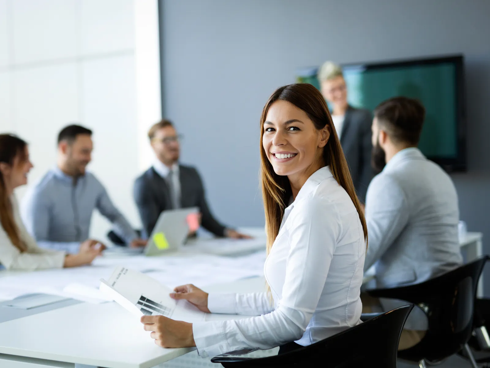 Smiling businesswoman holding documents during a meeting with diverse colleagues in a bright office conference room.