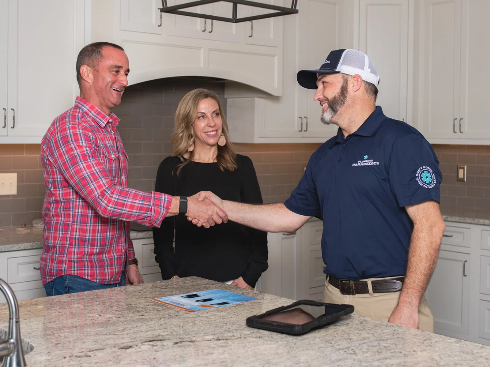 Paramedic shaking hands with man in kitchen with woman smiling nearby, tablet and brochure on granite countertop