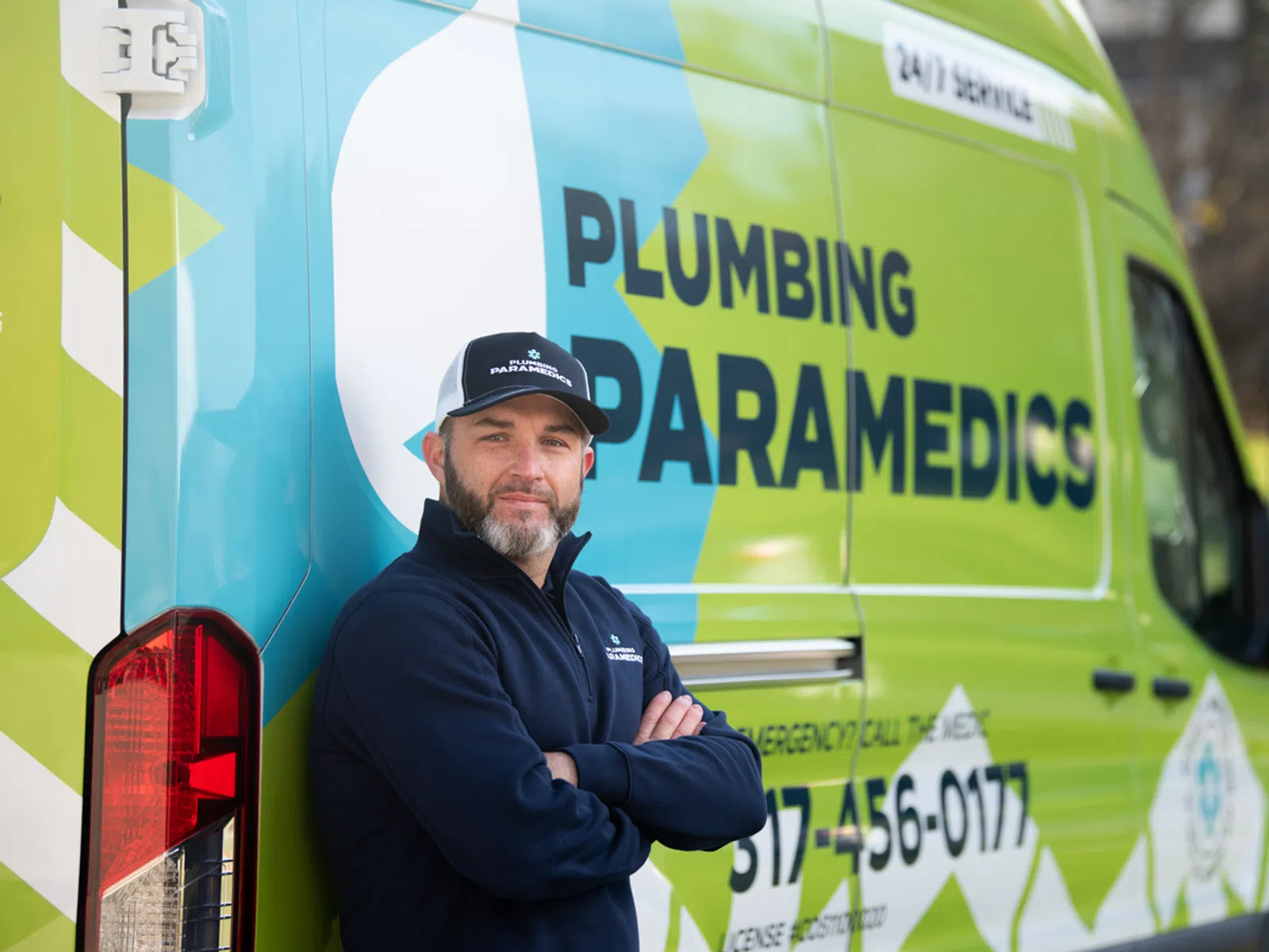Confident plumber in branded cap and jacket stands with arms crossed near Plumbing Paramedics service van