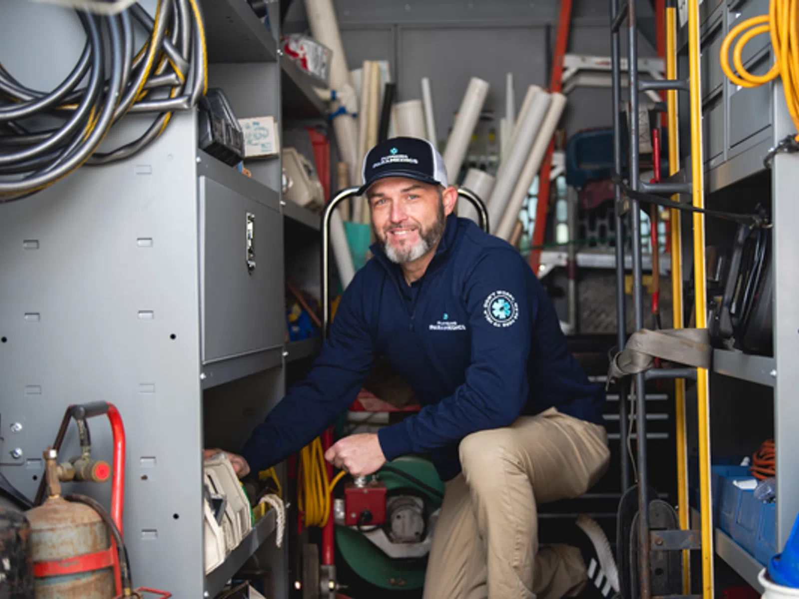 Smiling man in work uniform kneels inside a utility van surrounded by tools, hoses, and equipment.