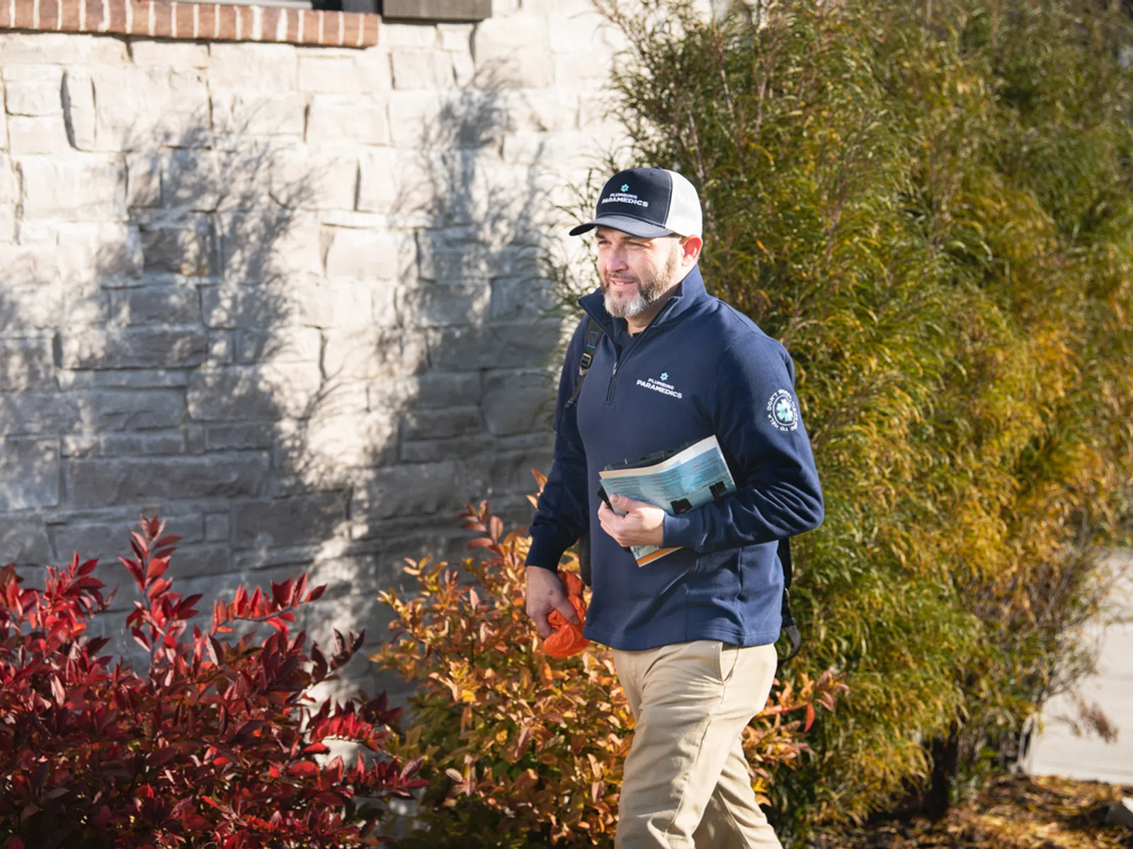 Man in navy jacket and cap walking outdoors near stone wall and colorful autumn bushes holding a newspaper.