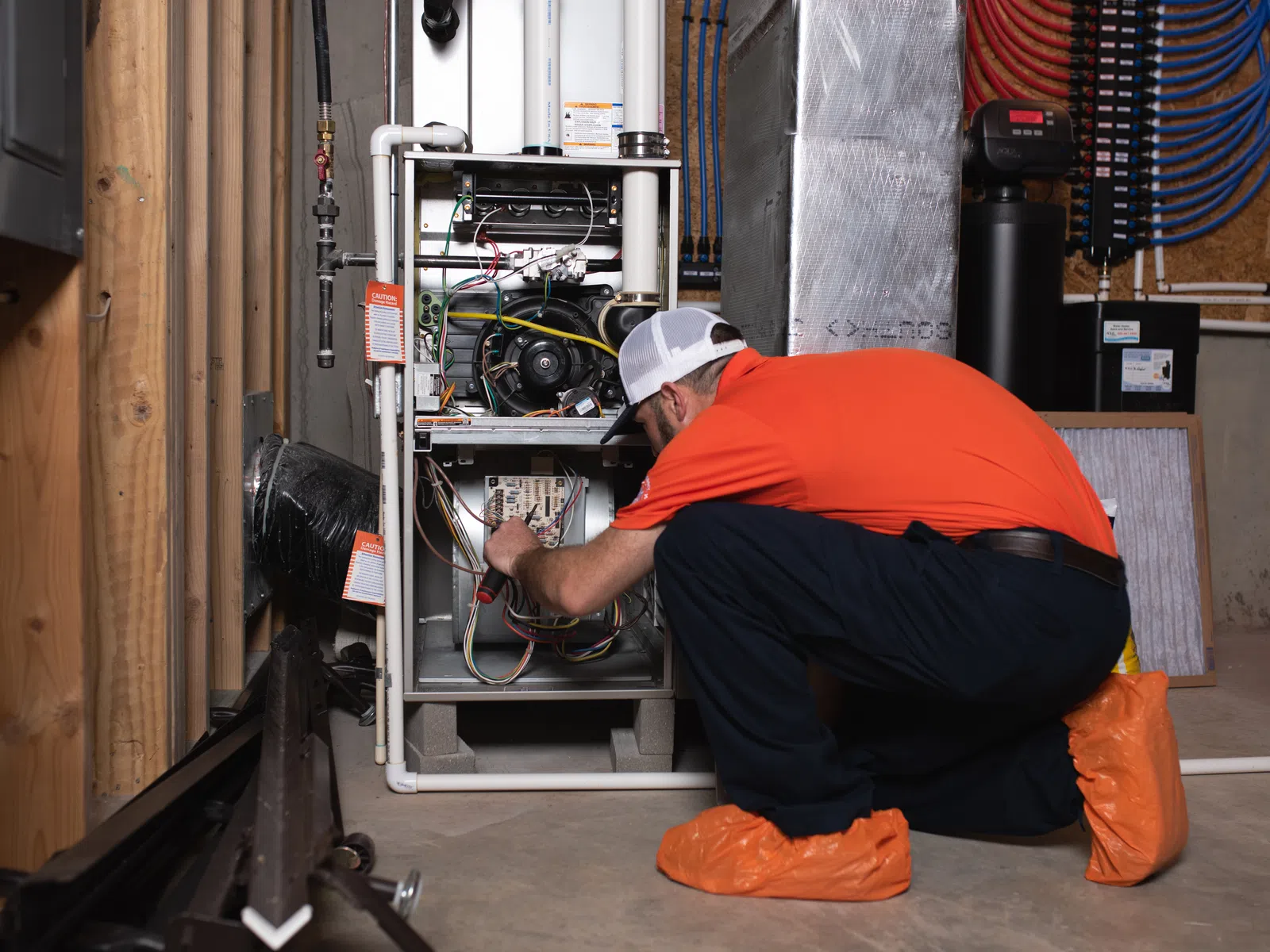 Technician in orange shirt repairing furnace system in basement wearing protective shoe covers and using a screwdriver.
