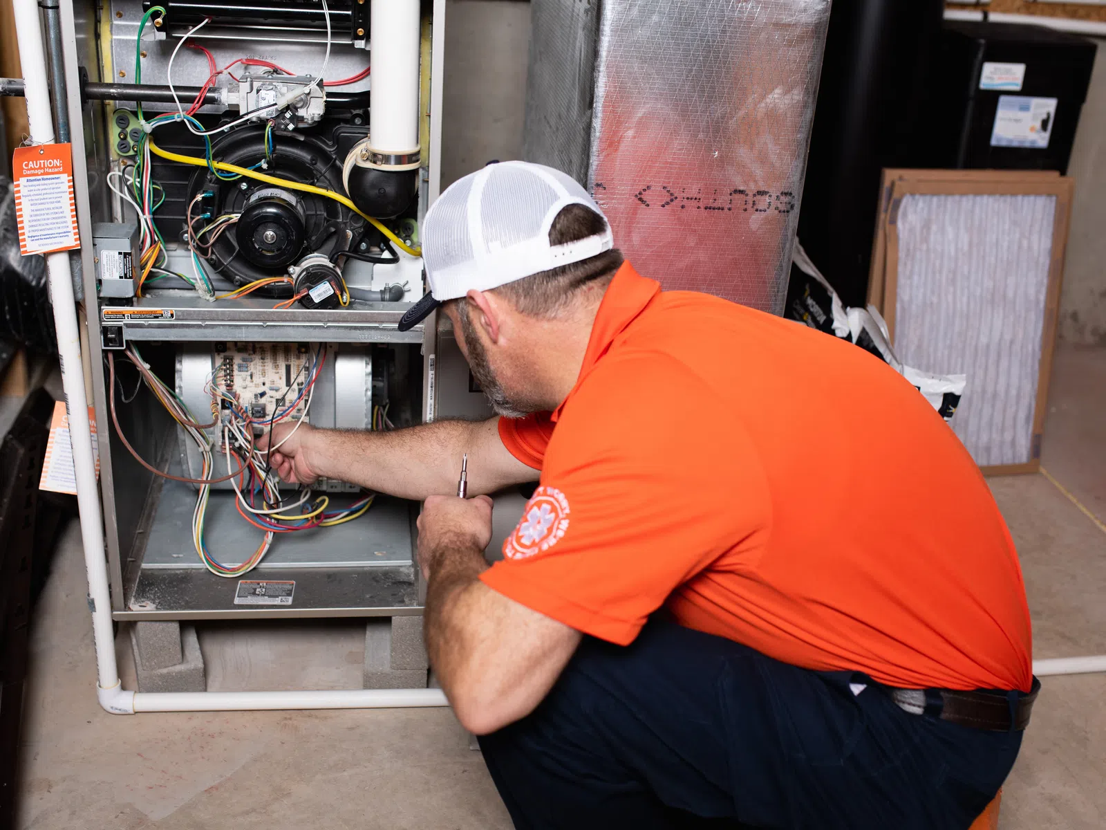 Technician in orange shirt inspecting and repairing wiring inside a furnace in a basement setting.