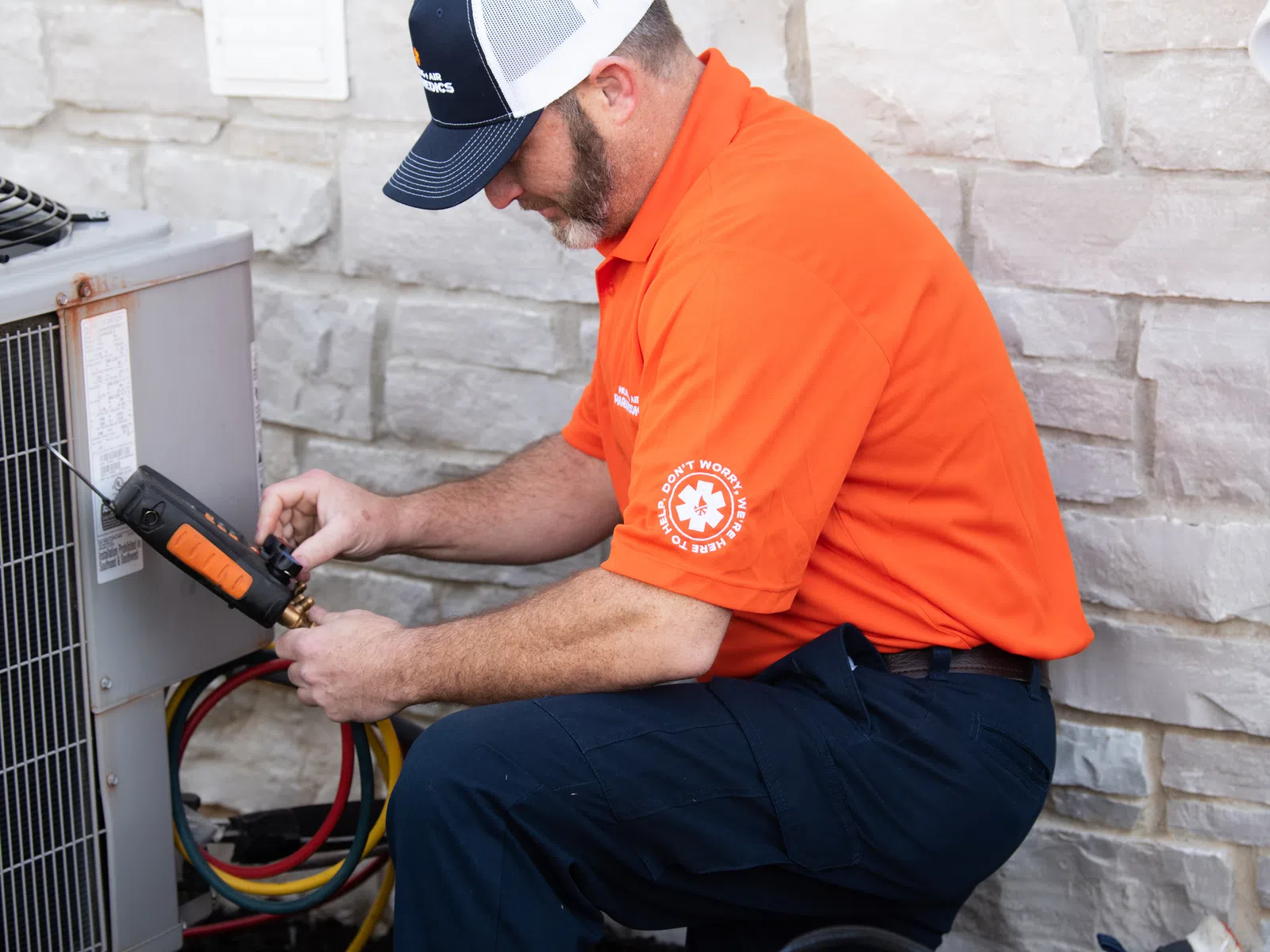 Technician in orange shirt and cap repairing outdoor HVAC unit against stone wall, kneeling with tools at work.