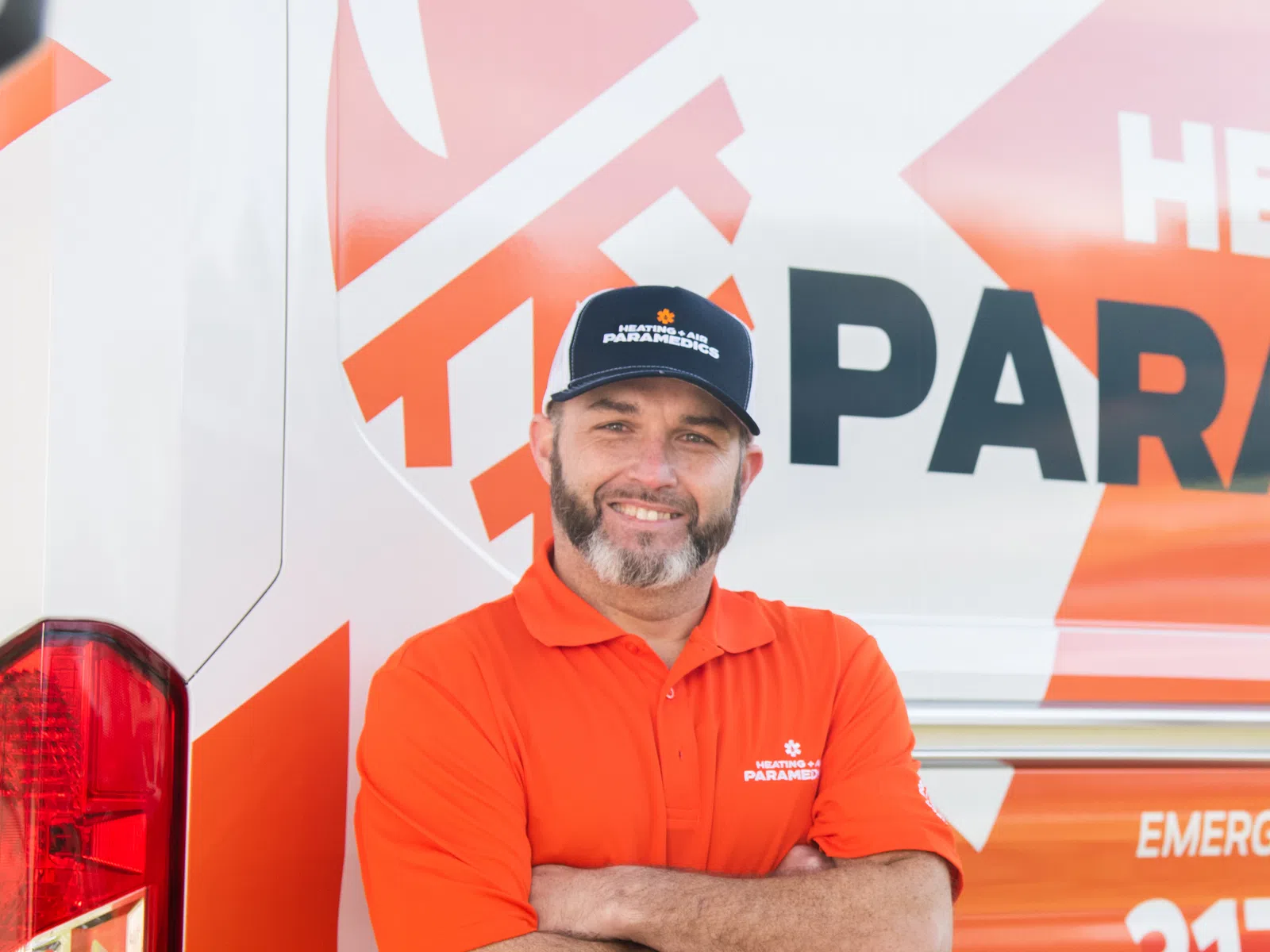 Smiling technician in orange shirt and cap stands with arms crossed by company service van with bold orange and white branding.