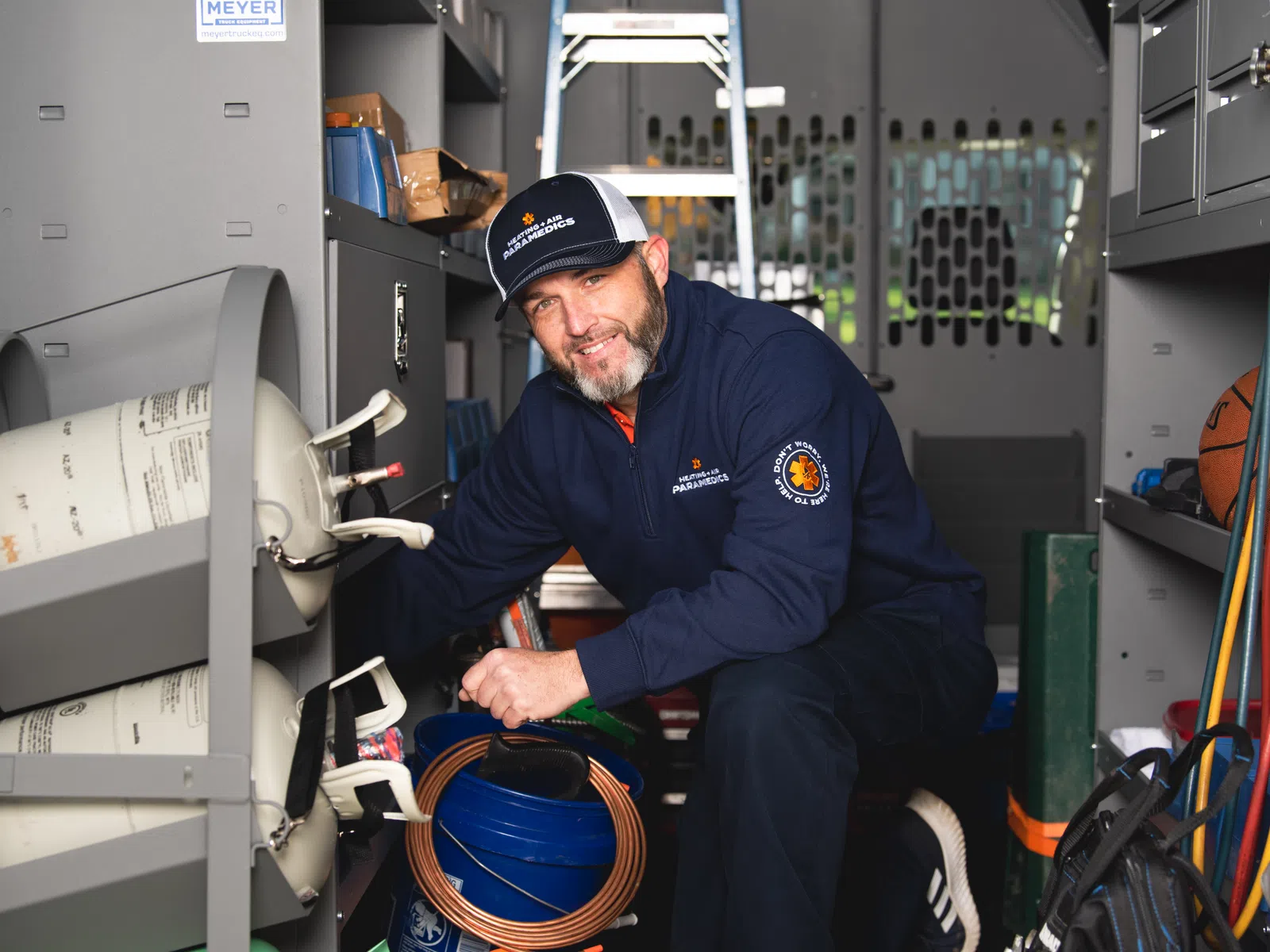 Male technician in uniform kneeling in a utility van organizing tools and gas cylinders for HVAC work
