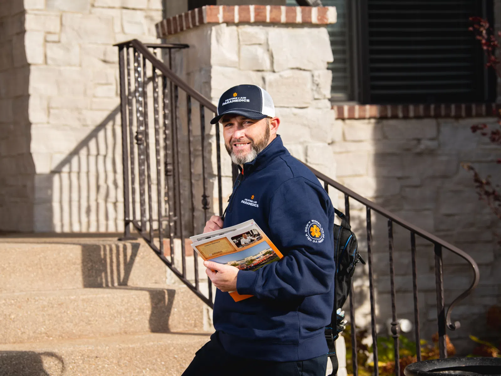 Smiling man in paramedics uniform holding brochures standing on house steps in daylight.
