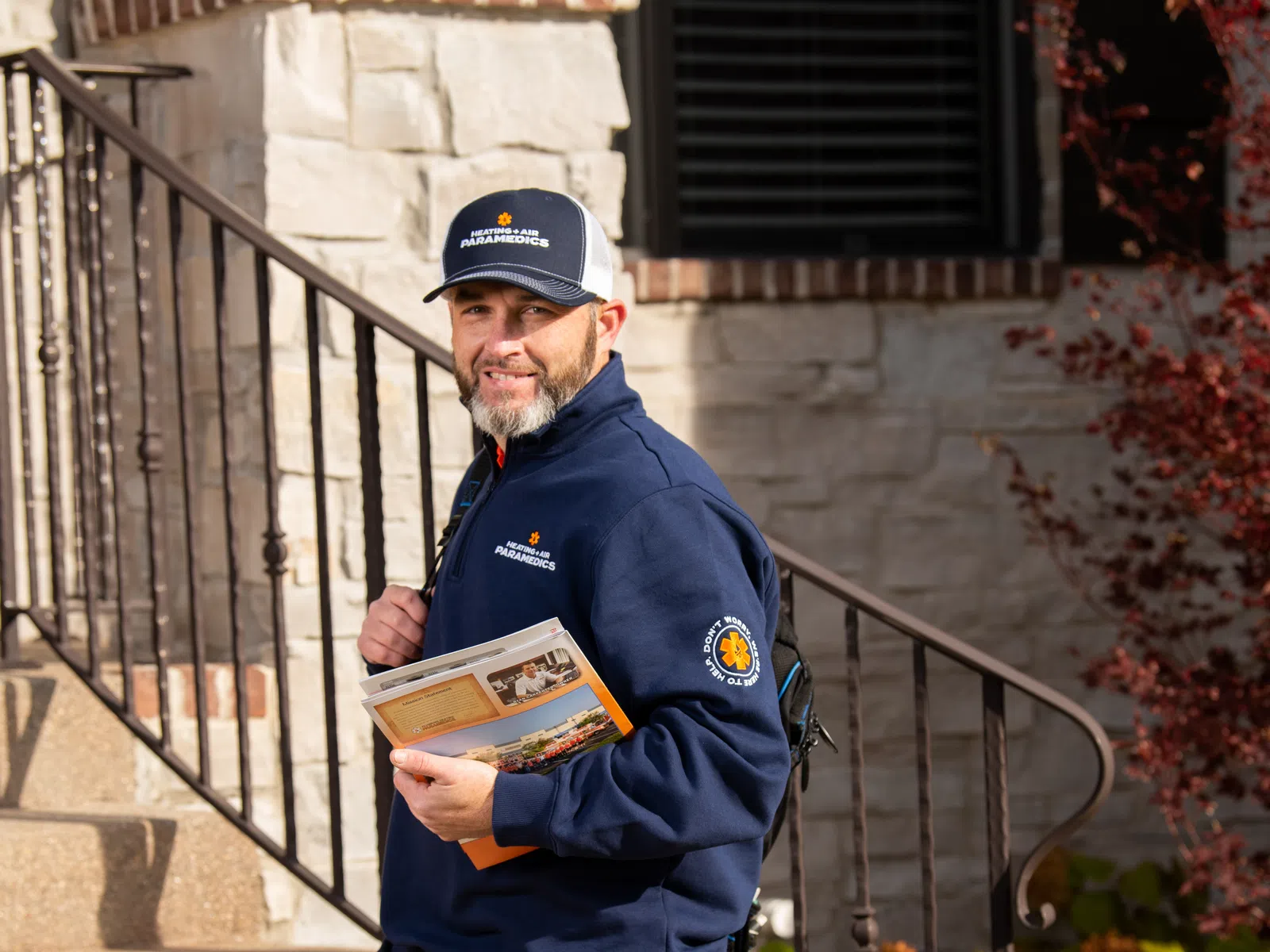 Paramedic in navy blue uniform holding a booklet, standing on outdoor stairs by a stone building and railing.
