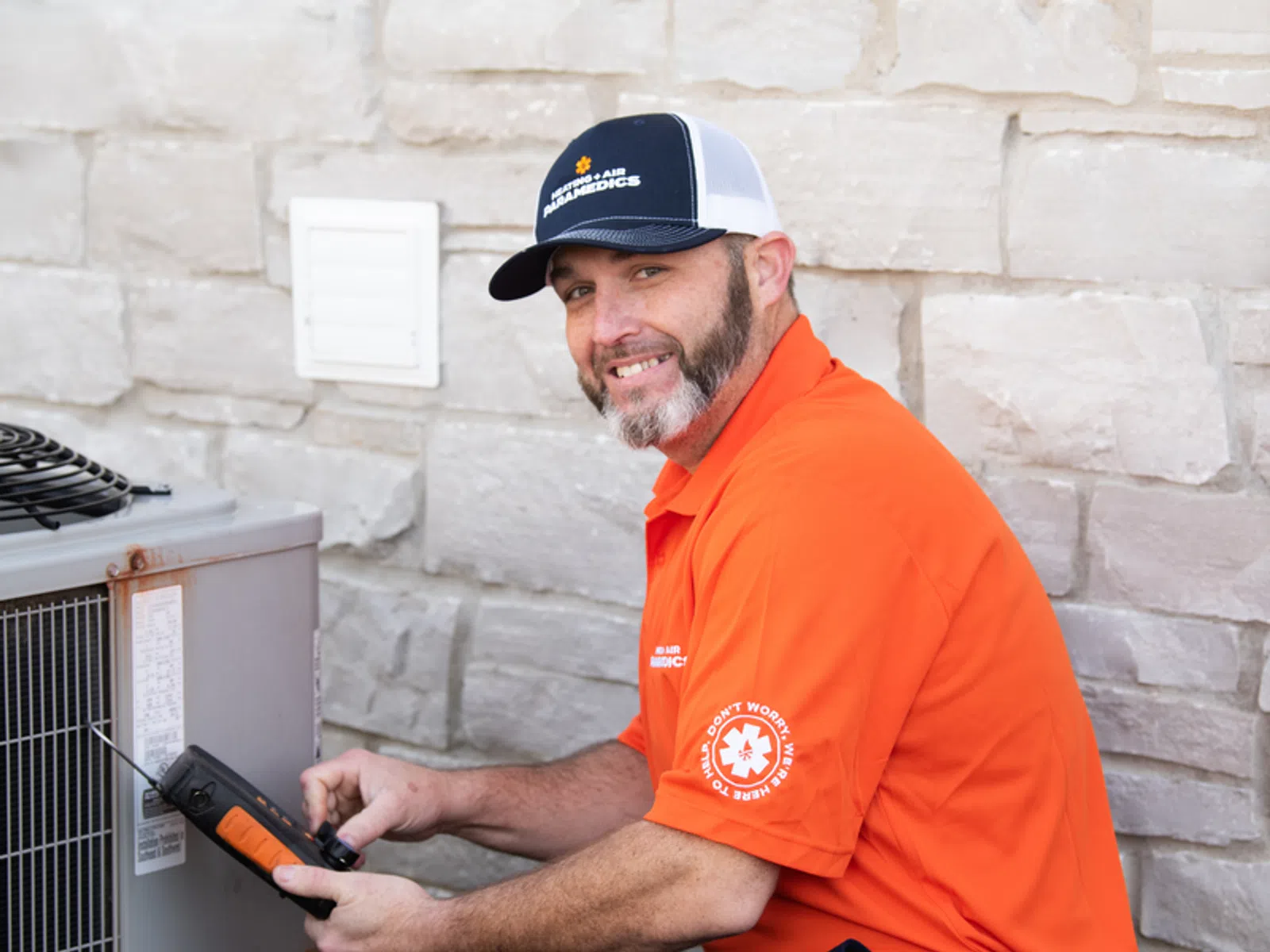 Technician in orange shirt repairing an outdoor HVAC unit against a stone wall background.