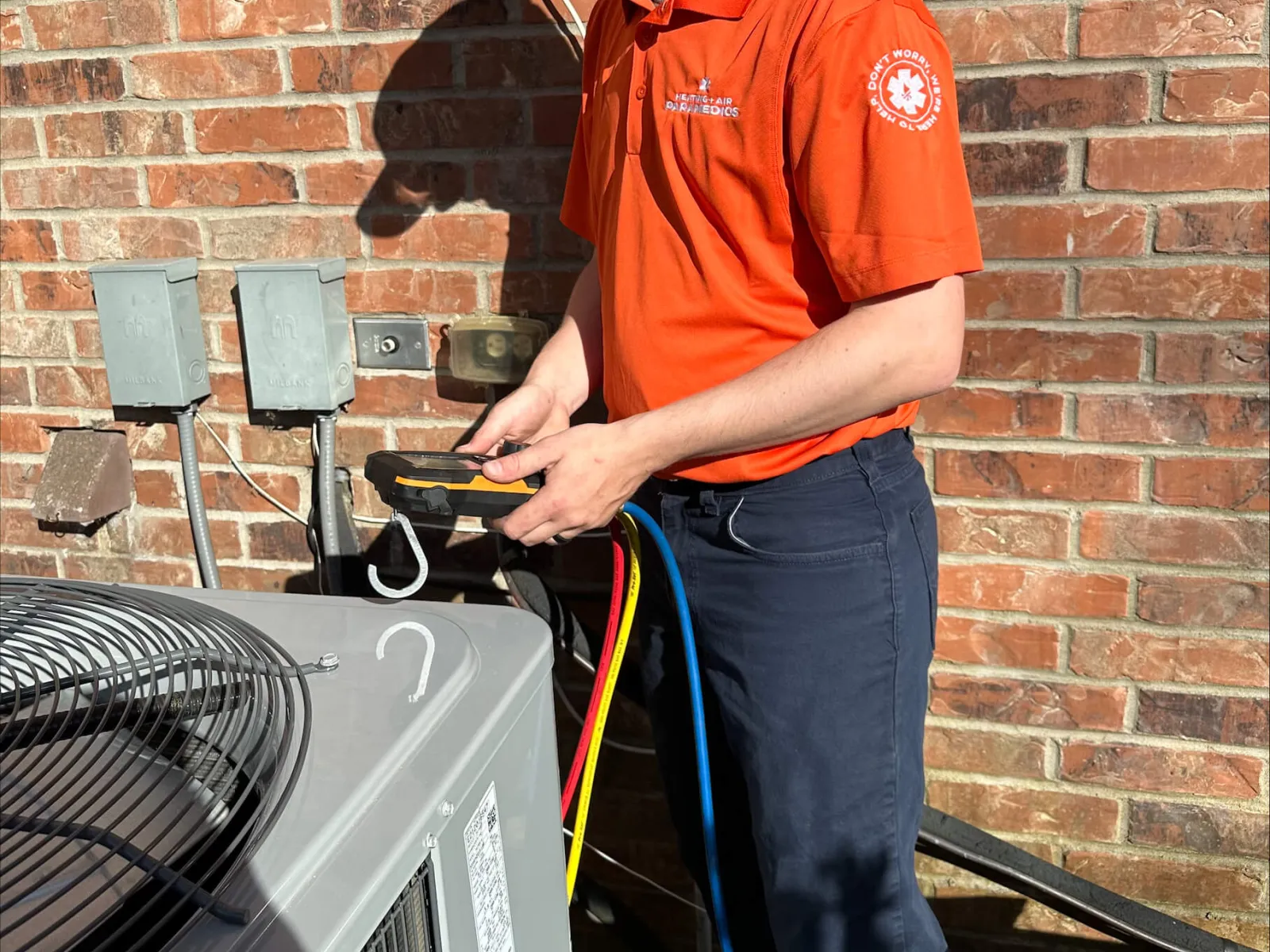 Technician in orange shirt and cap checking outdoor HVAC unit with diagnostic tools against a brick wall background