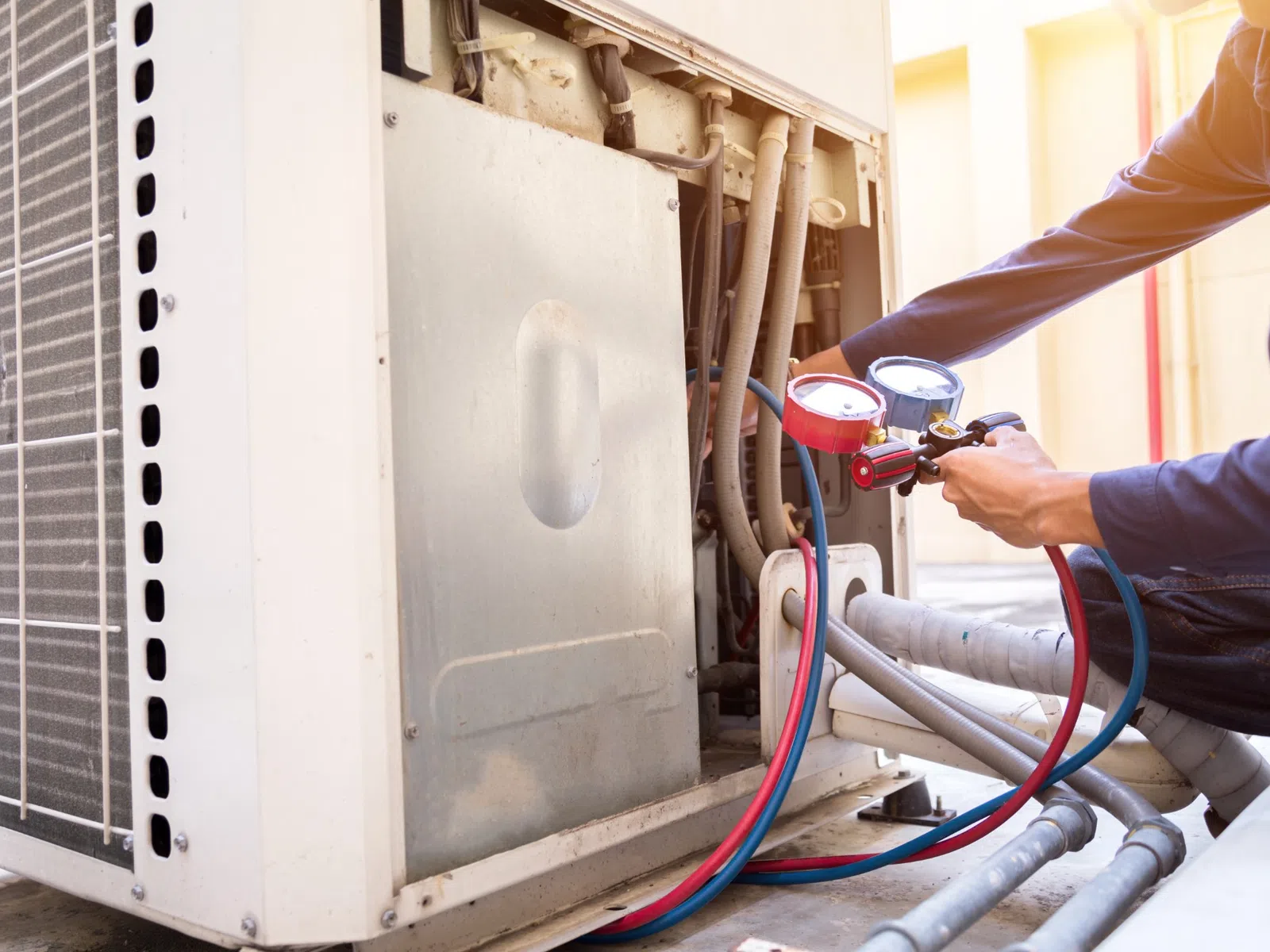 Technician adjusts gauges on outdoor air conditioning unit with red and blue hoses connected for maintenance.