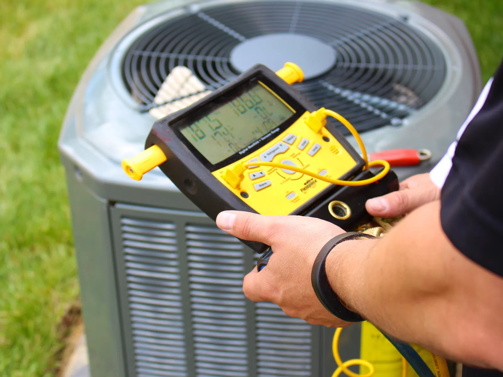 Technician using a yellow digital HVAC gauge to check air conditioning unit performance outdoors on green grass.