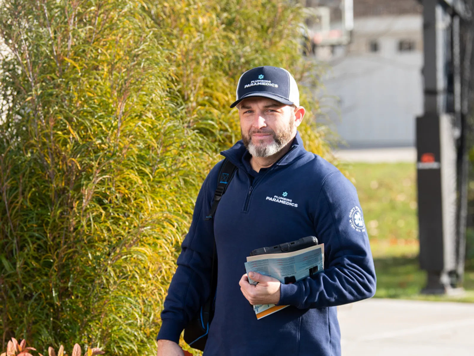 Male paramedic in navy jacket and cap holding papers outdoors near basketball hoop and autumn foliage.