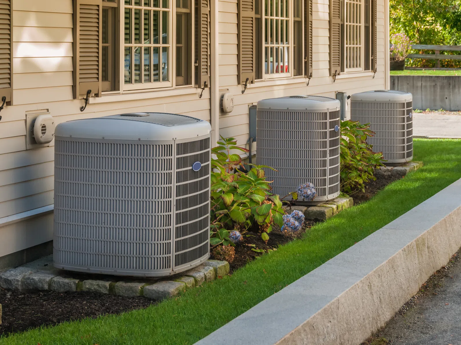 Three outdoor air conditioning units installed along the side of a beige house with green grass and plants.